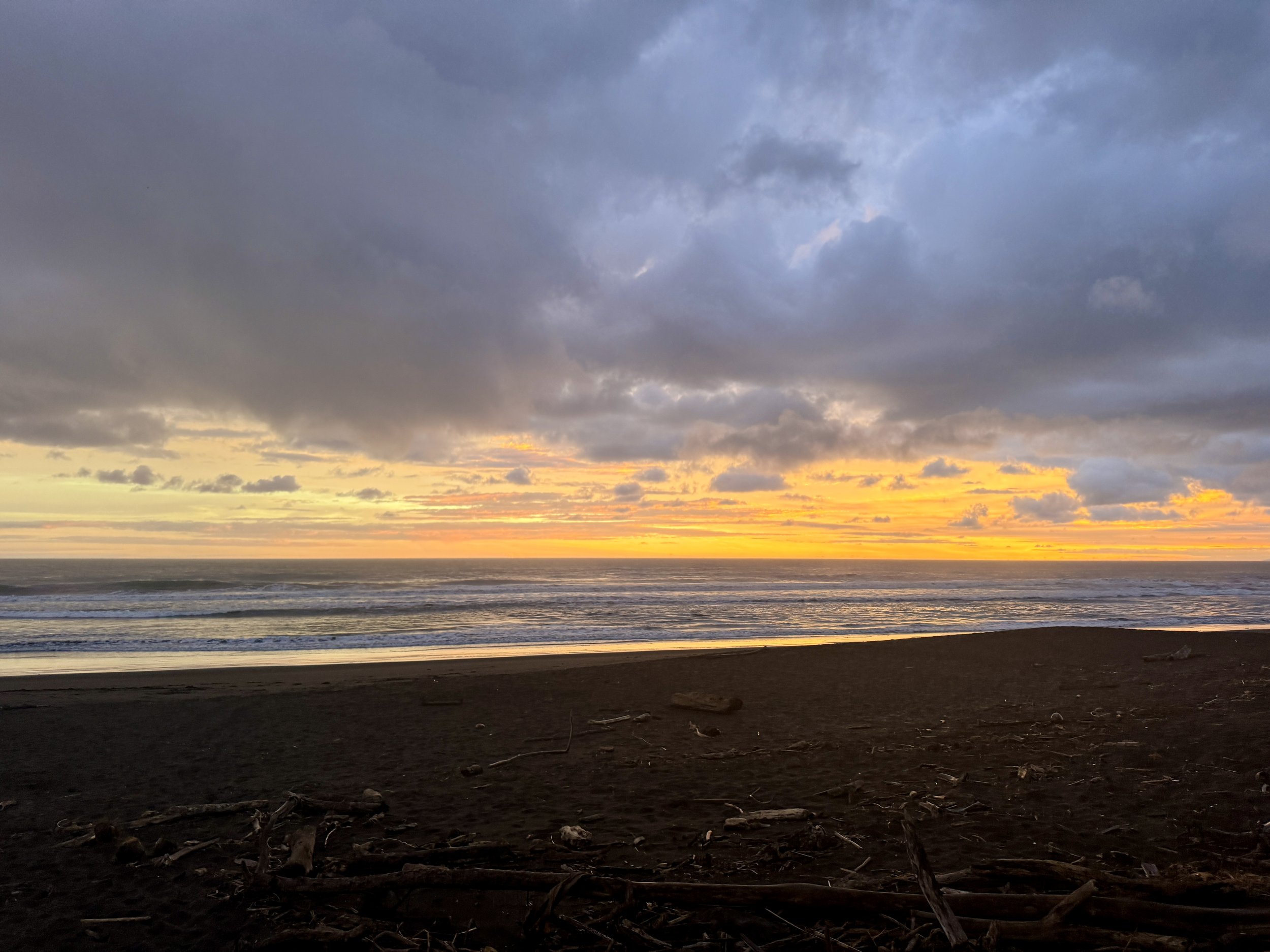 Golden sunset over Hermosa Beach with dark sand in the foreground and dramatic storm clouds above the ocean.