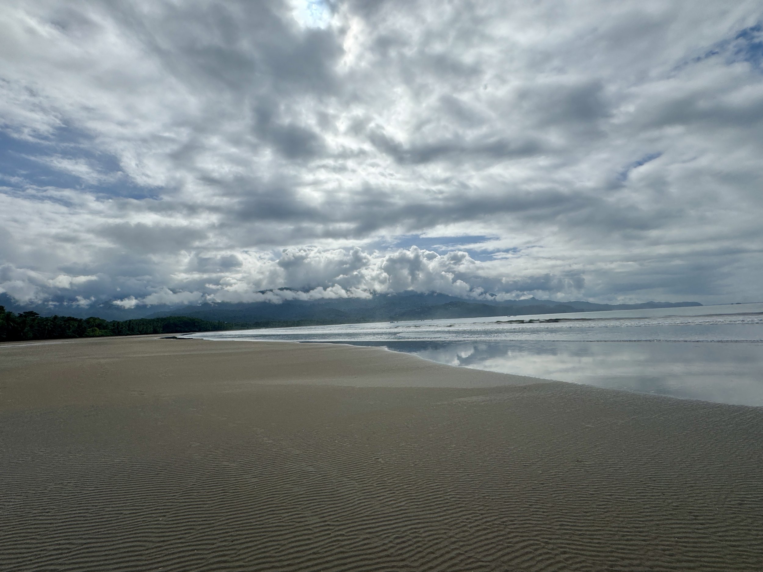 Wide sandy beach at low tide with dramatic cloud cover and mountains in the distance at Uvita.