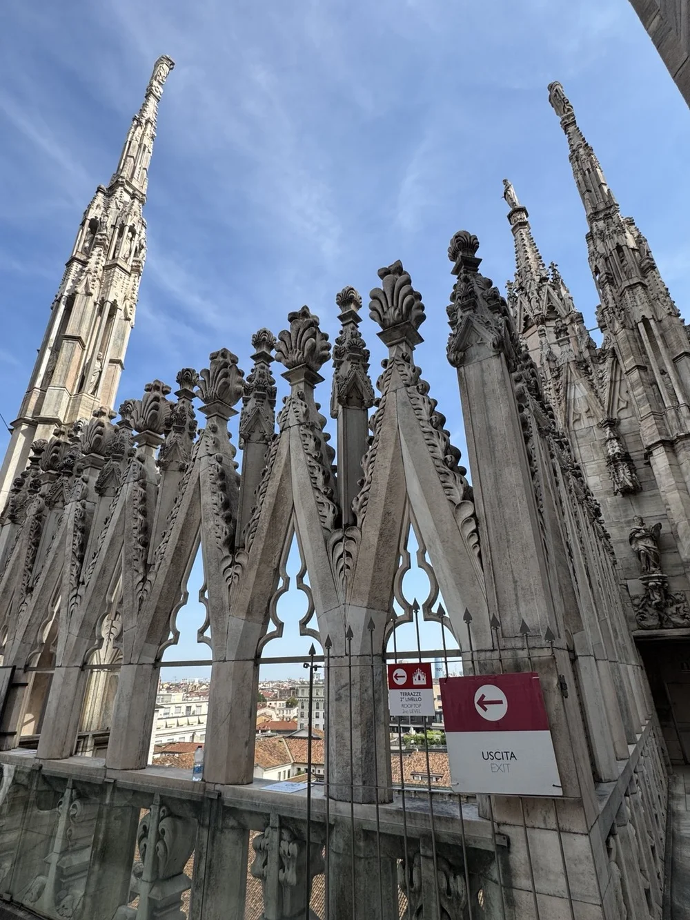 Ornate marble spires and rooftop walkway on the Duomo di Milano terraces under a blue sky.