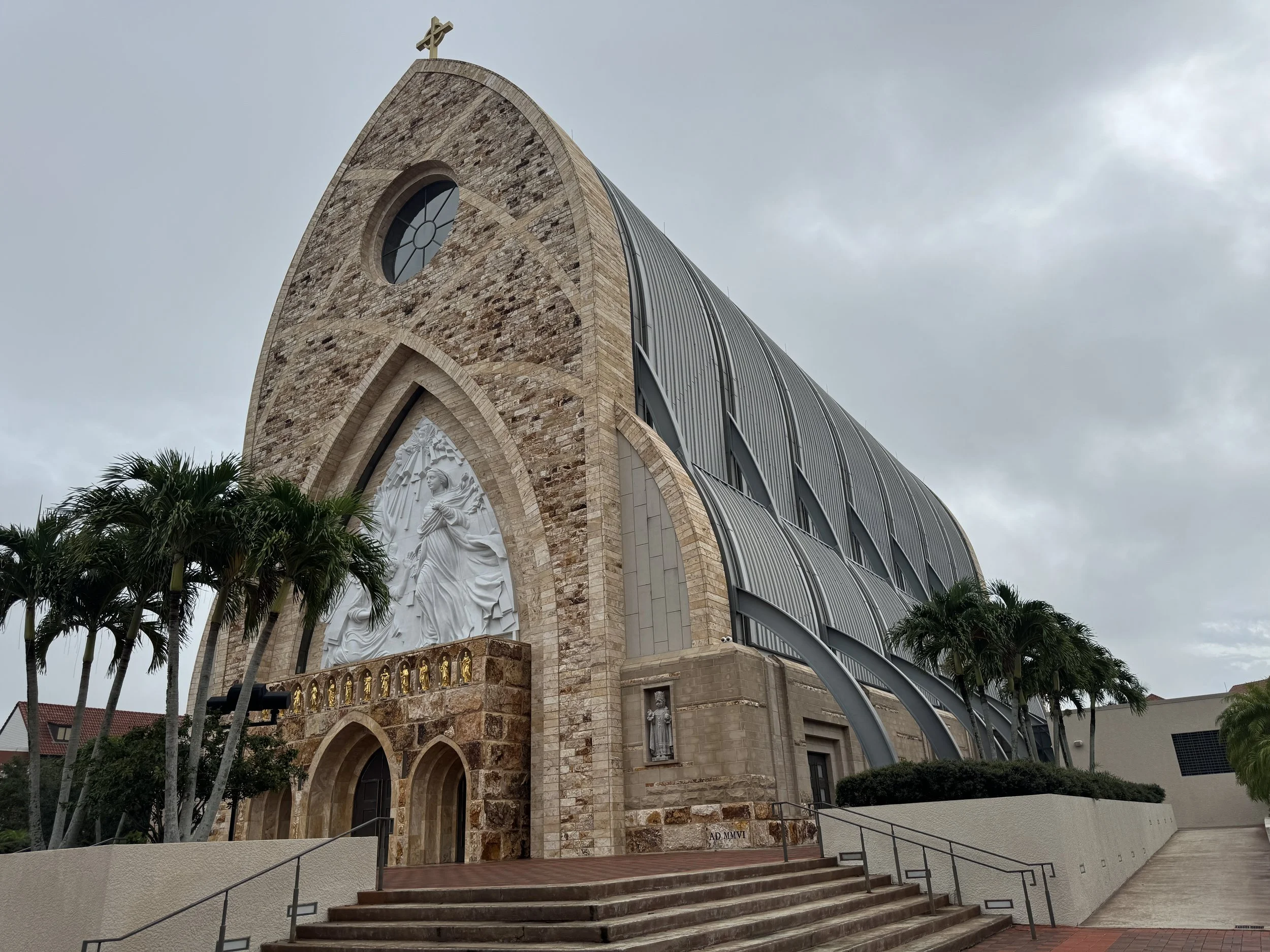 Side perspective of the large Ave Maria Catholic Parish Church showing the tall arched architecture and palm trees surrounding the entrance.