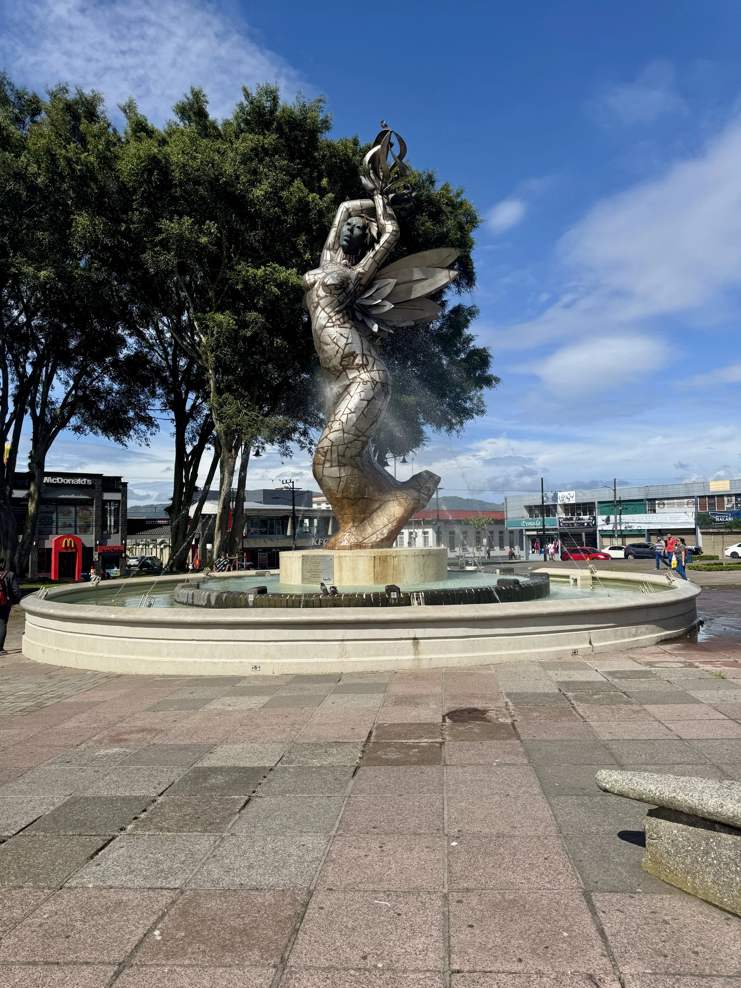 Fountain sculpture in Cartago plaza near the ruins with palm trees and bright blue sky.