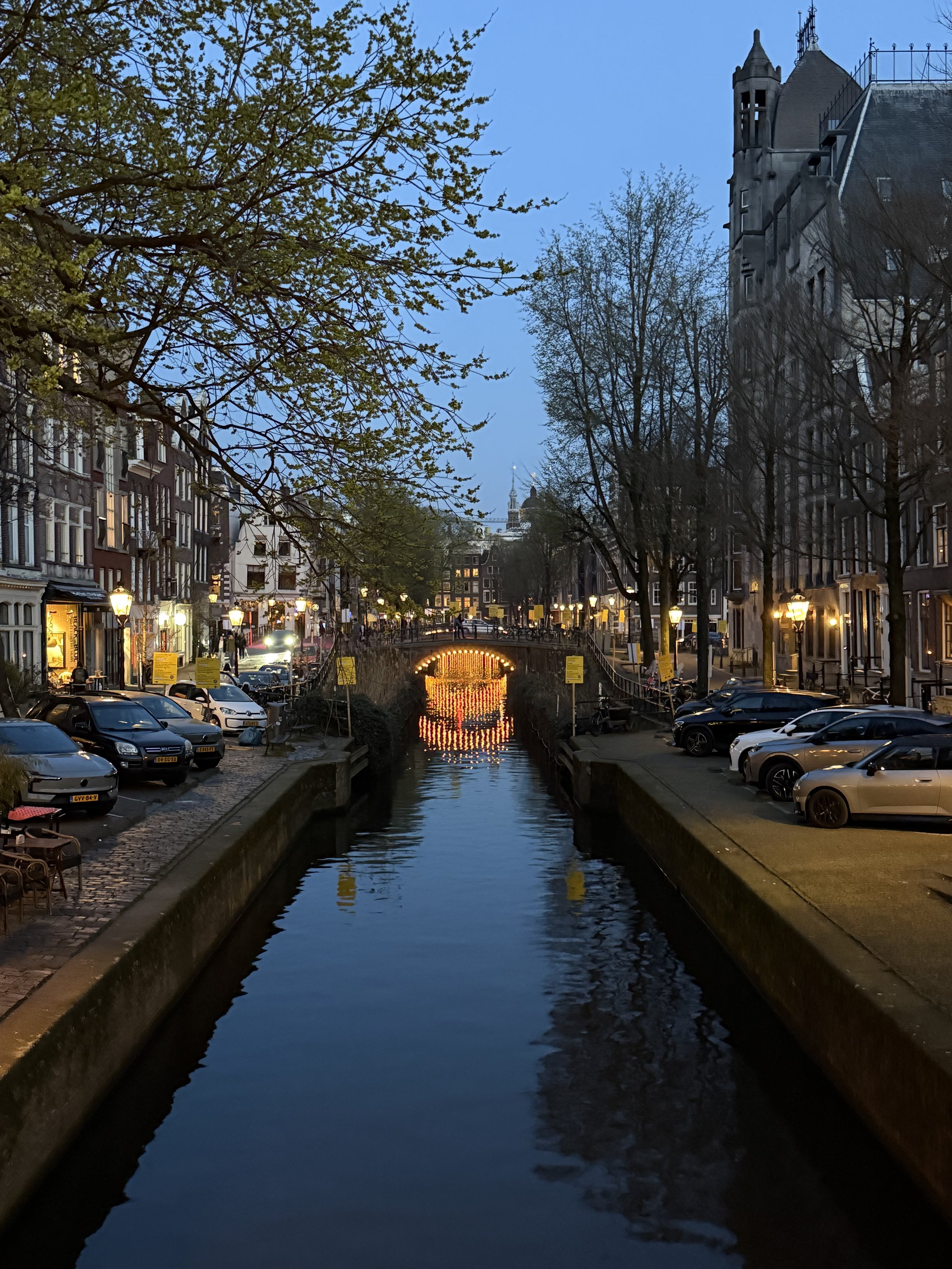 Evening view of an Amsterdam canal with glowing lights reflecting on the water and trees lining the street