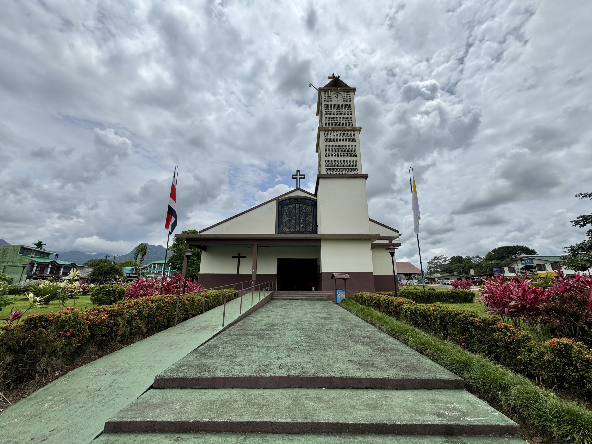Iglesia Católica de La Fortuna with walkway leading through the park under a cloudy sky.