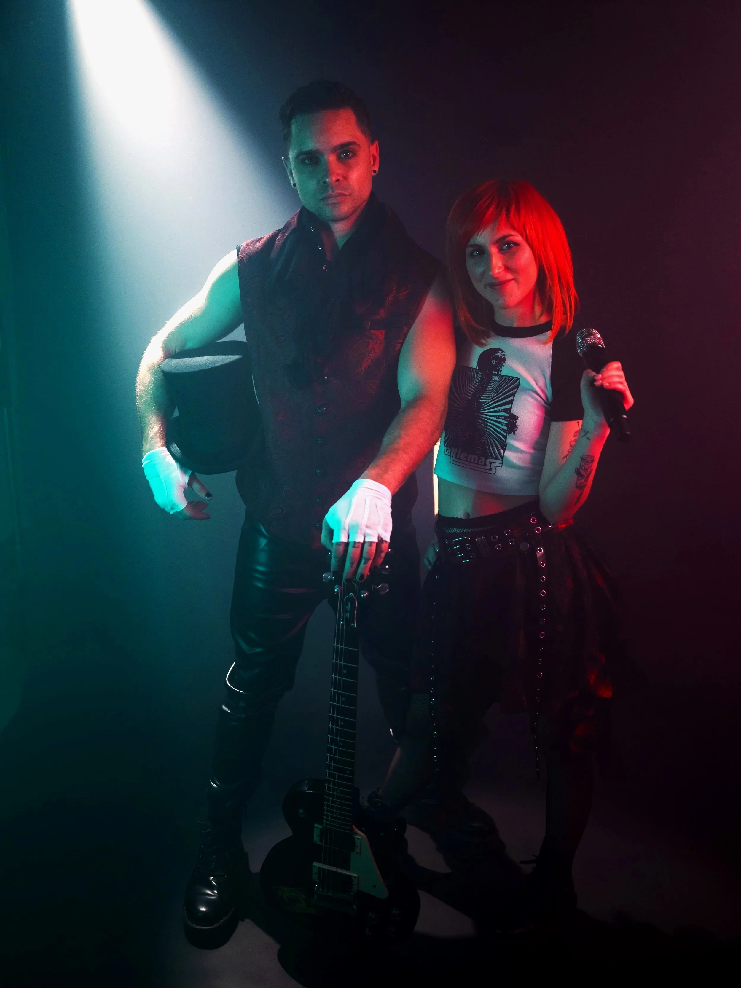 Image of a man and a woman standing next to each other in a spot light with clothing reminiscent of Paramore and Panic! At the Disco