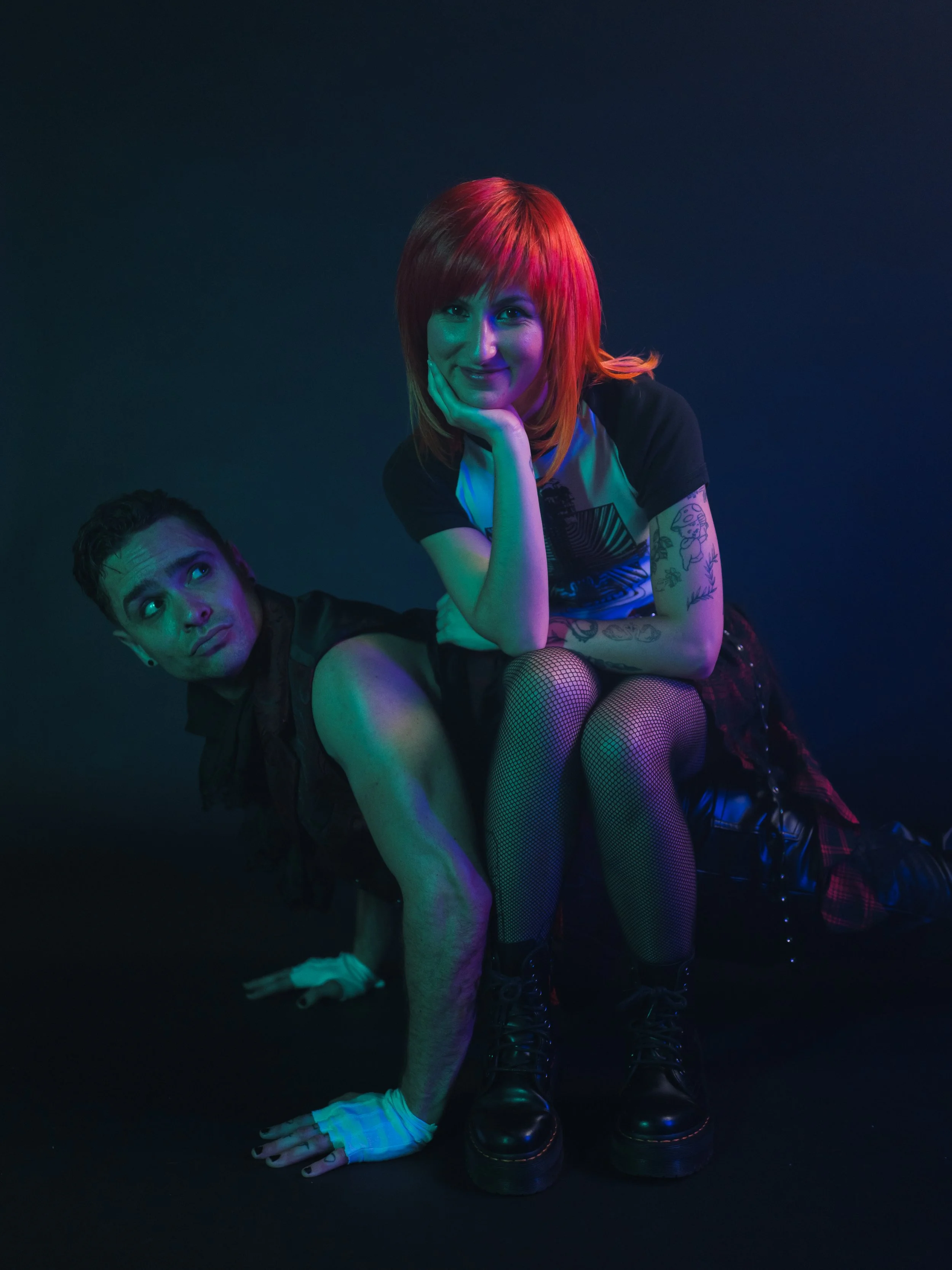 A woman with bright red hair, wearing a black t-shirt with a graphic, fishnet tights, and black boots, sitting on a man. The man has dark curly hair, is wearing a sleeveless black shirt, has tattoos on his arms, and is on all fours on the floor. They are lit with colorful, stage-like lighting against a dark background.