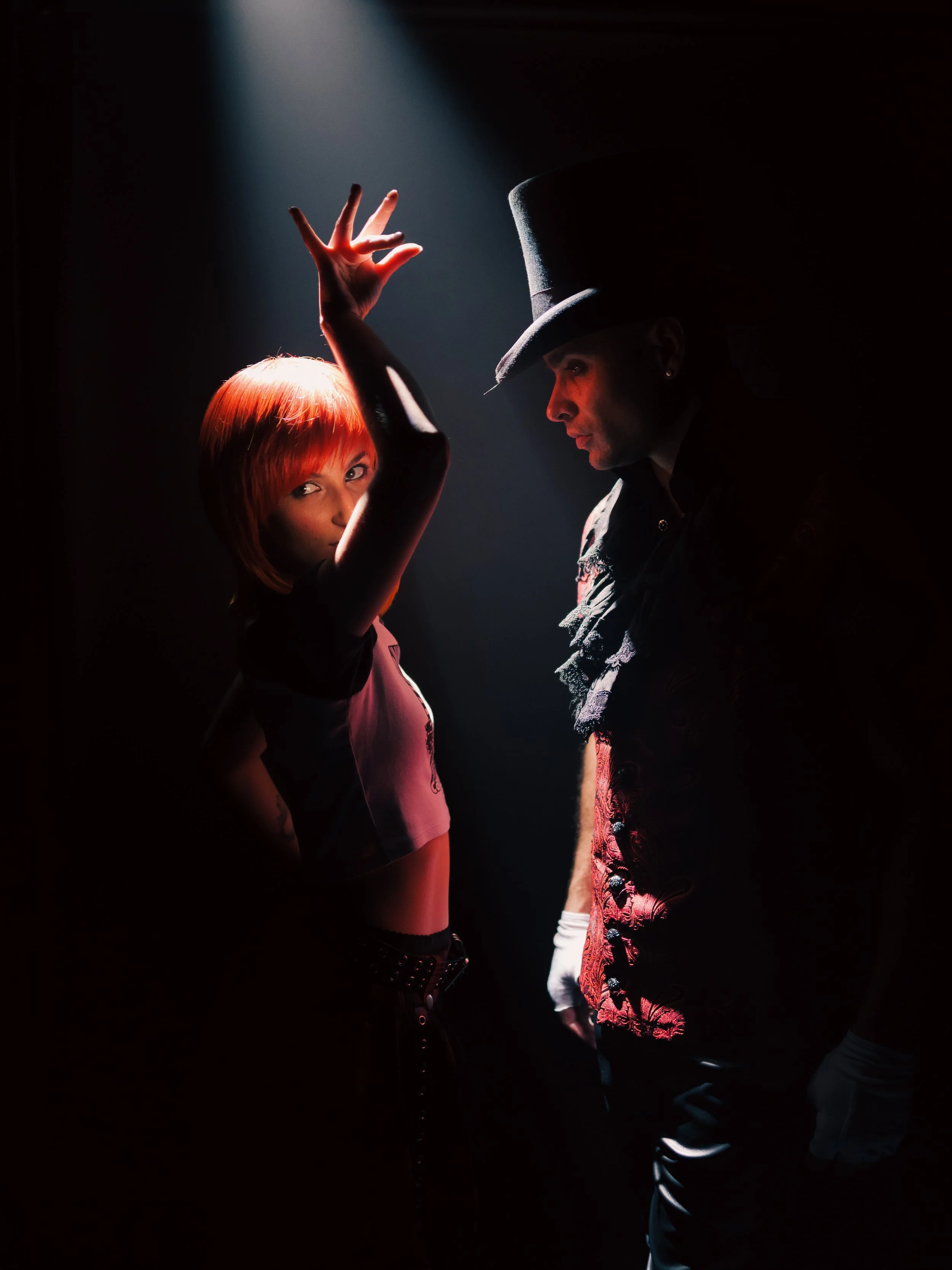 A woman with bright red hair and a man wearing a tall top hat and dark clothing facing each other in a dramatic pose under a spotlight in a dark background.