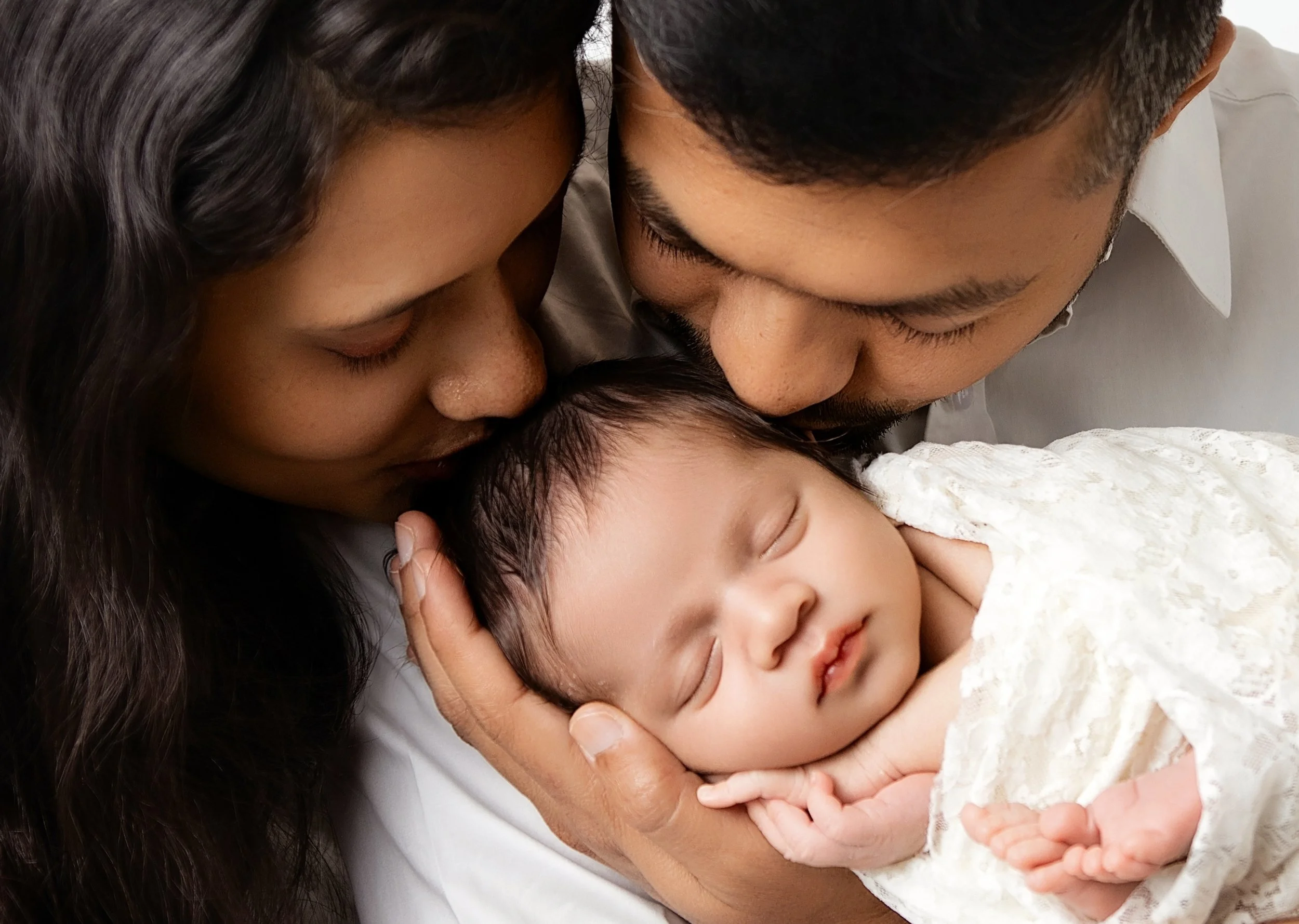 A family of three holding their sleeping newborn baby, with the mother and father close to the baby's face, showing a tender moment.