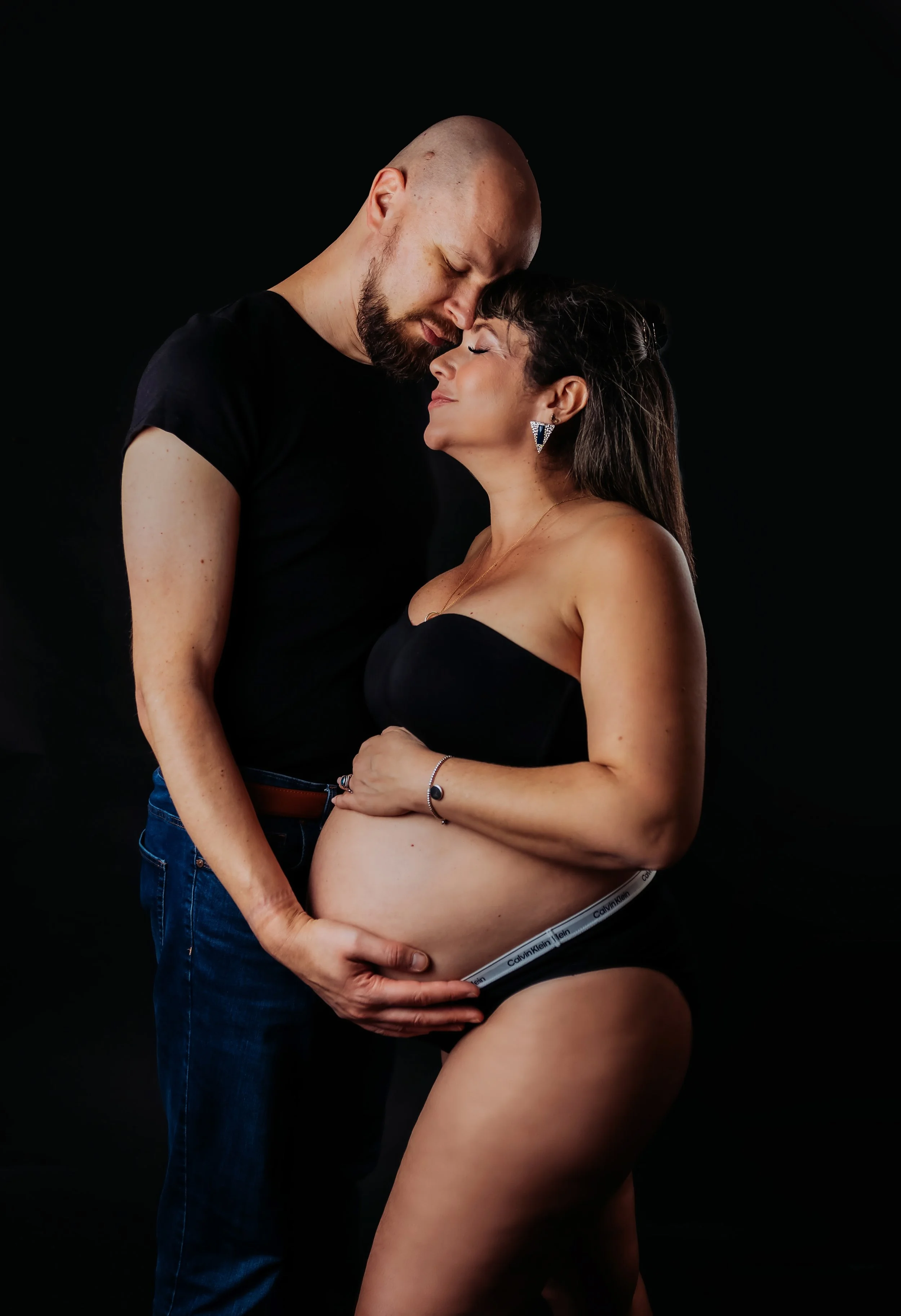 a dark portrait of a couple. the pregnant lady Is infront wearing a black bra with her husband infront of her with his head against hers. his in a black t shirt cradling her bump. backdrop is black