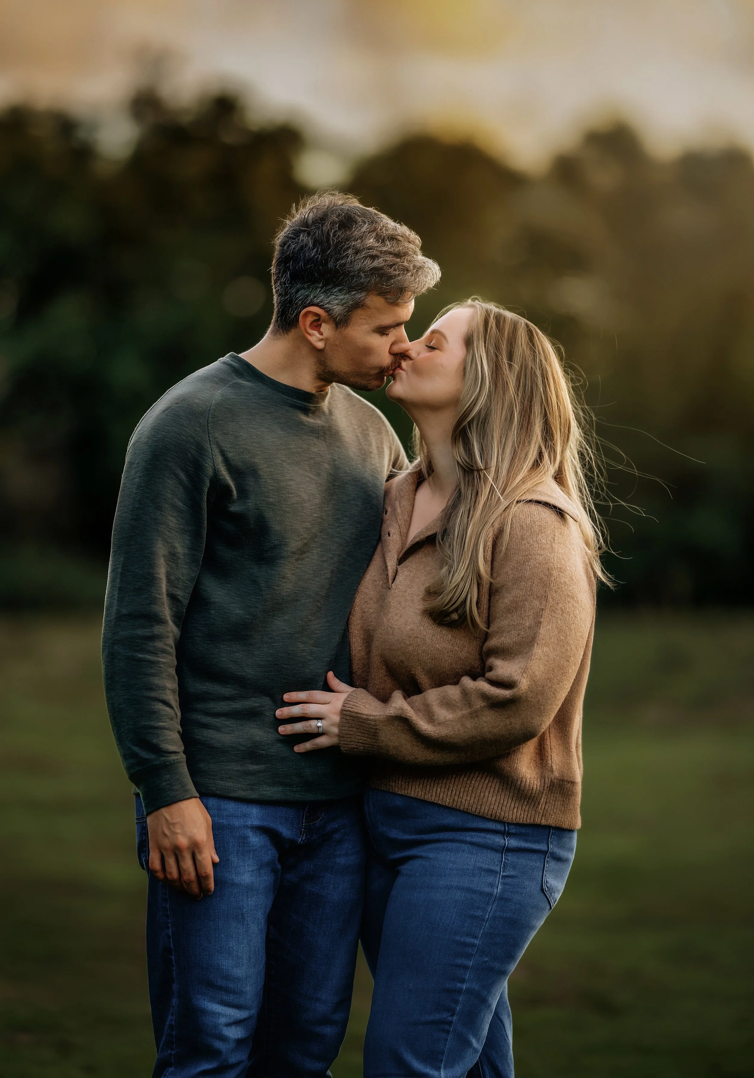 A couple kissing outdoors at sunset, with green trees in the background.