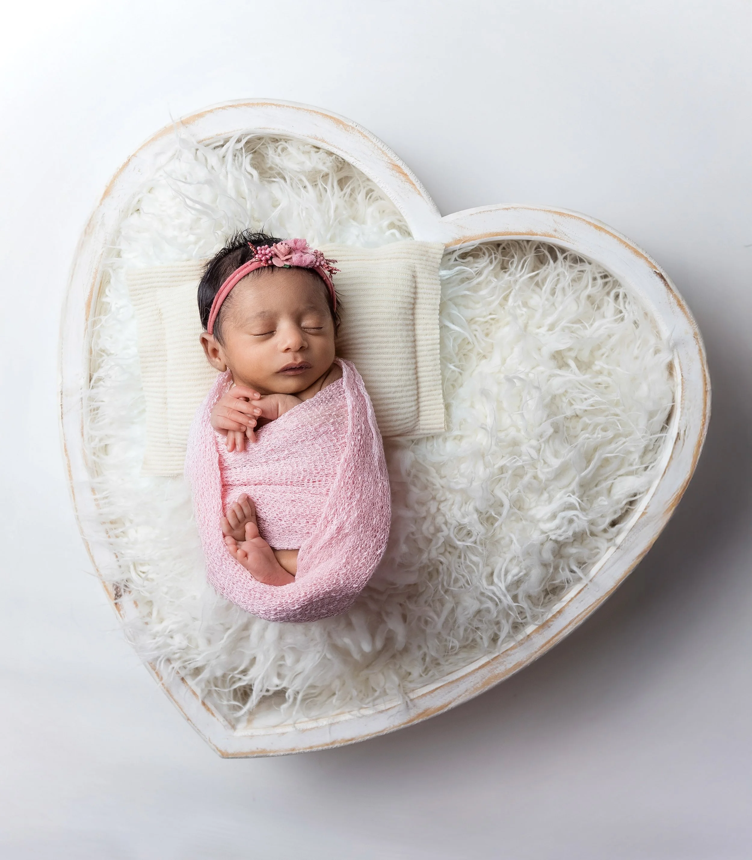 newborn girl in a pink wrap with a pink headband laying in a heart filled with a fluffy white blanket In a white background