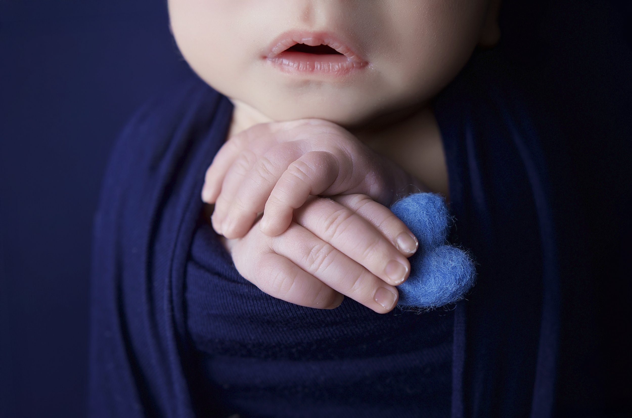 Close-up of a child's face with lips slightly parted, wearing a dark blue outfit. The child is holding a small piece of blue felt with one hand.