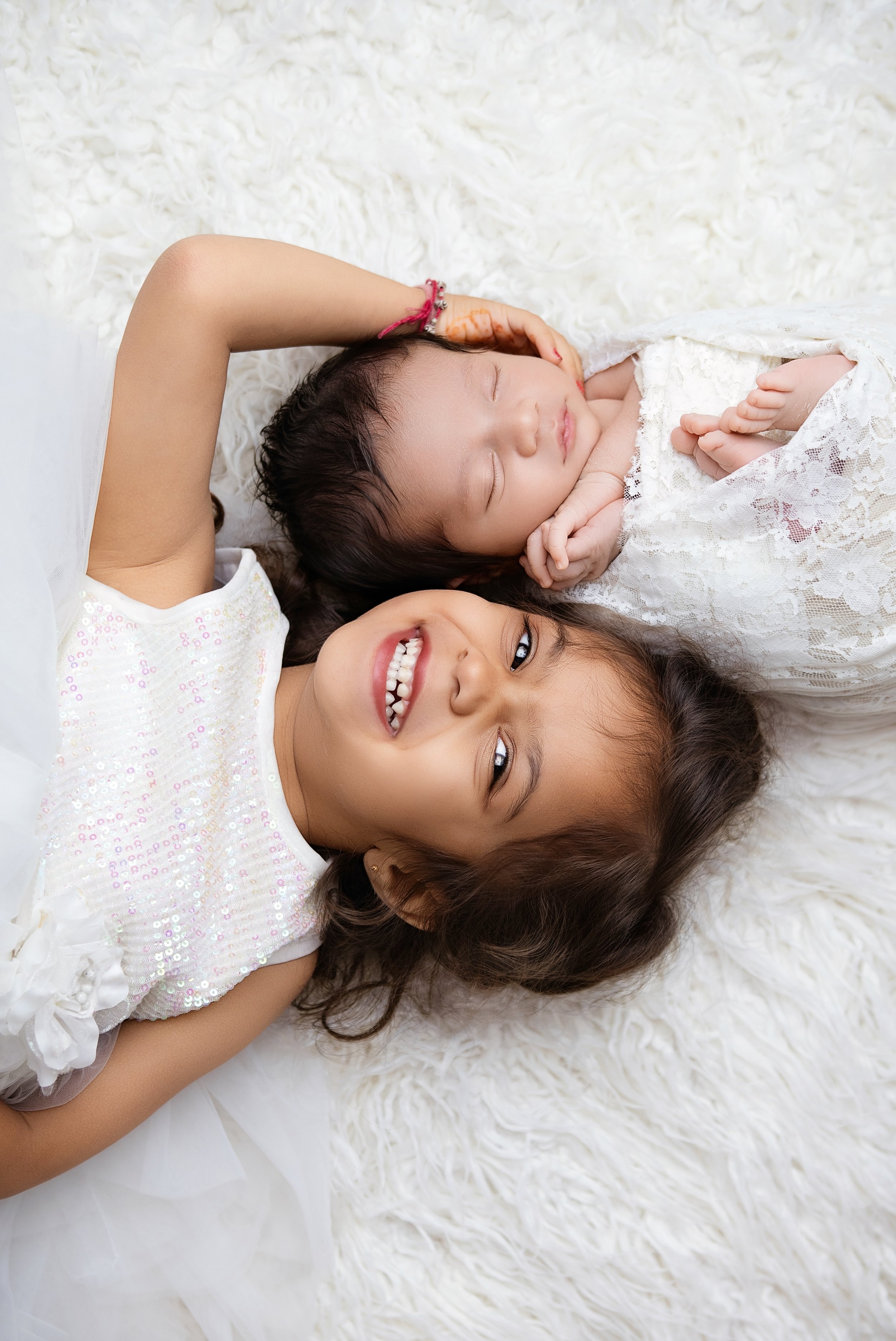 Two young girls lying on a plush, white surface, one smiling with a sparkle in her eyes and the other peacefully sleeping.