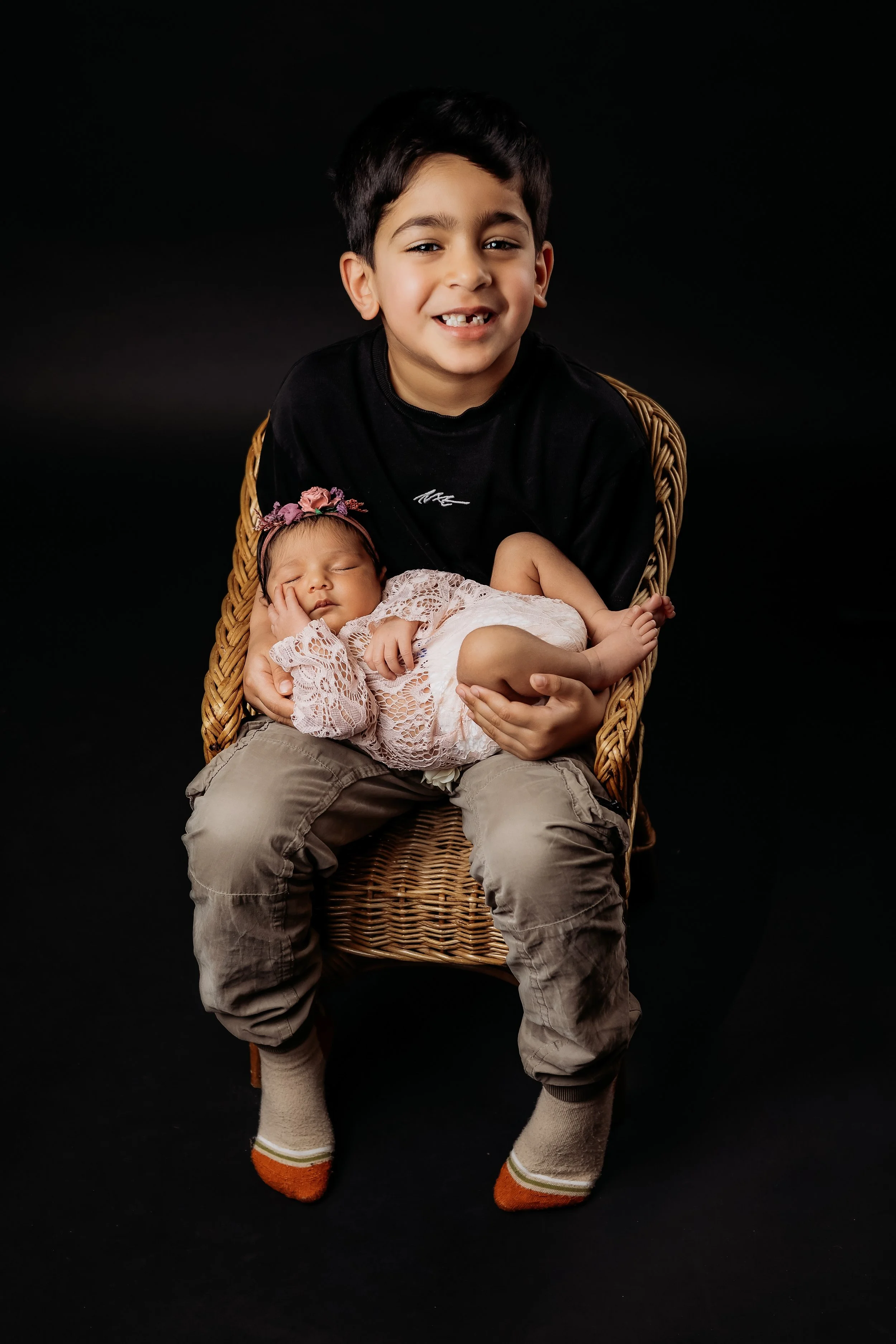 studio photo of boy sitting in wicker chair holding newborn sister. sister dressed in pink. boy is wearing black on a black backdrop