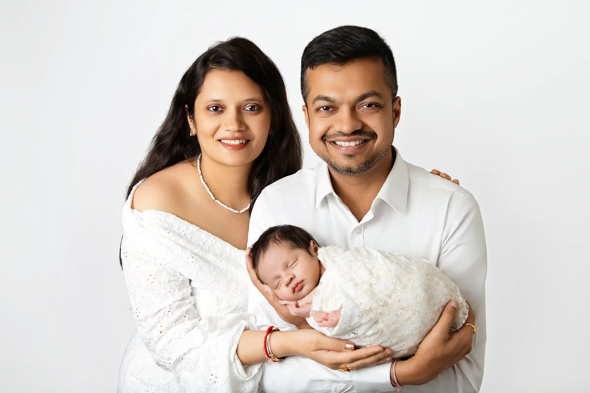 An Indian family of three, including a woman, man, and a newborn baby, smiling at the camera against a plain white background.