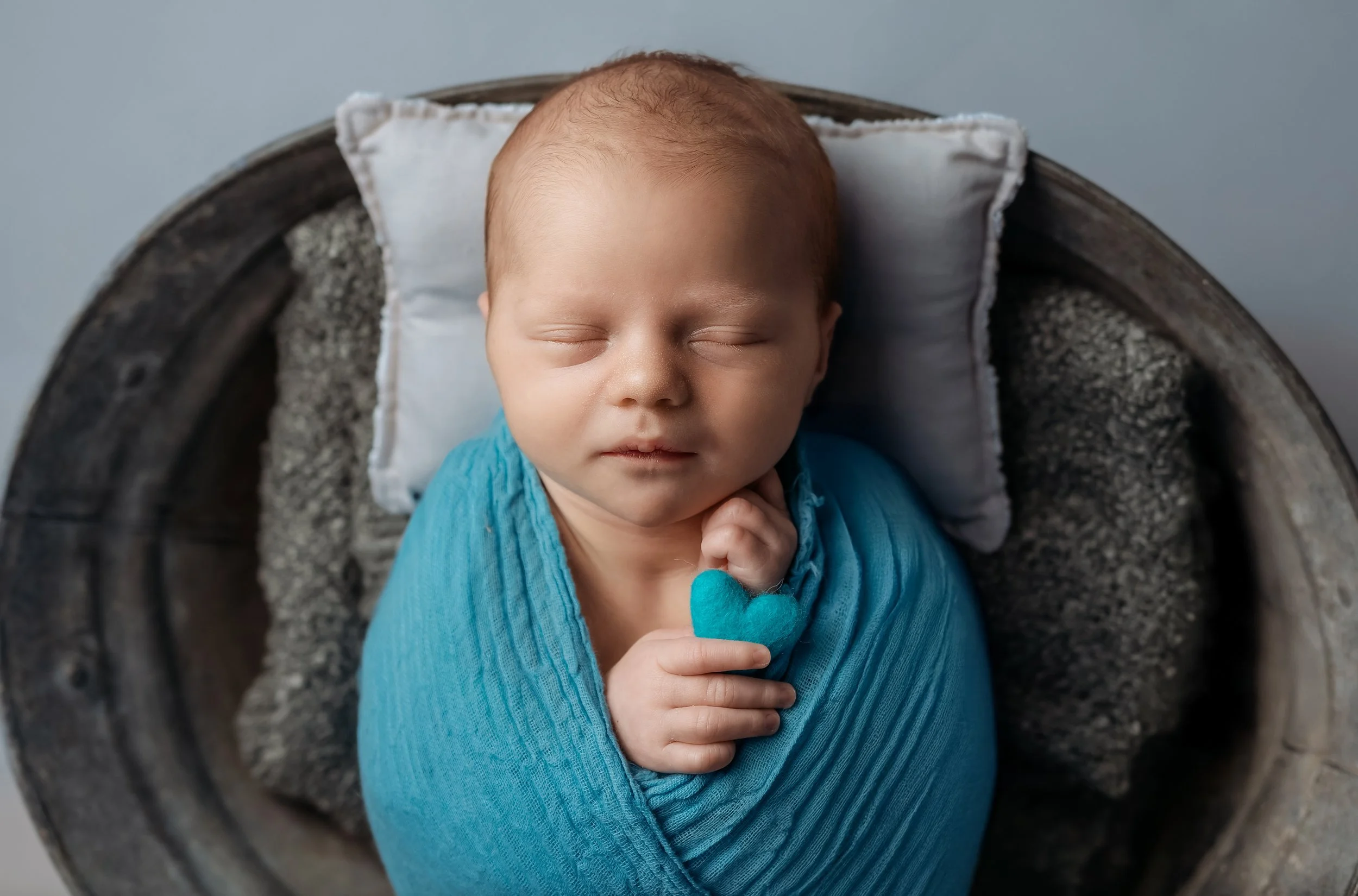 newborn boy with a blue wrap holding a blue felt heart on a blue pillow. laid on a grey posing bucket with a grey blanket. newborn studio photoshoot