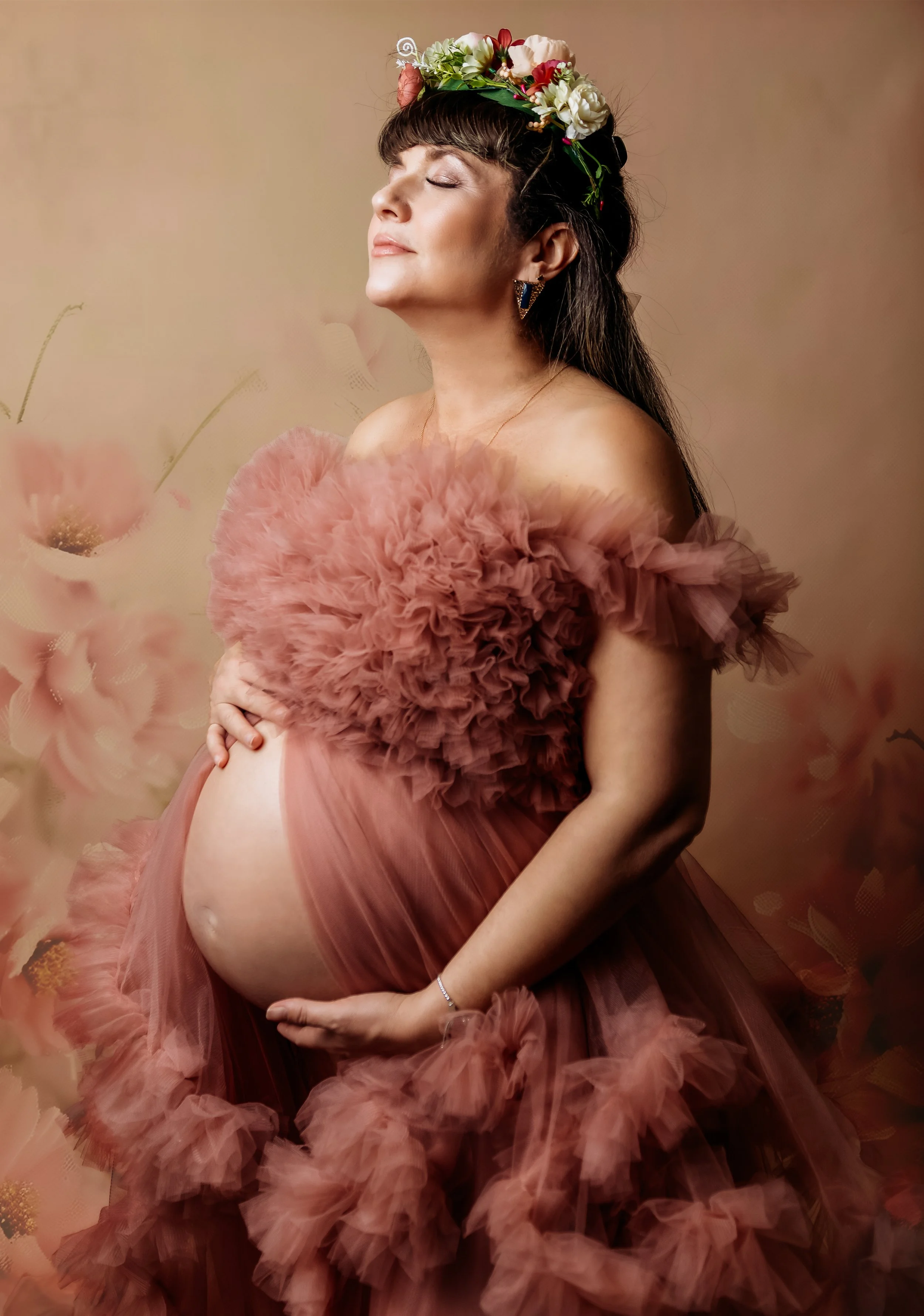A pregnant woman with a floral crown, wearing a pink ruffled dress, stands with her eyes closed and one hand on her belly against a soft blush background with floral elements.
