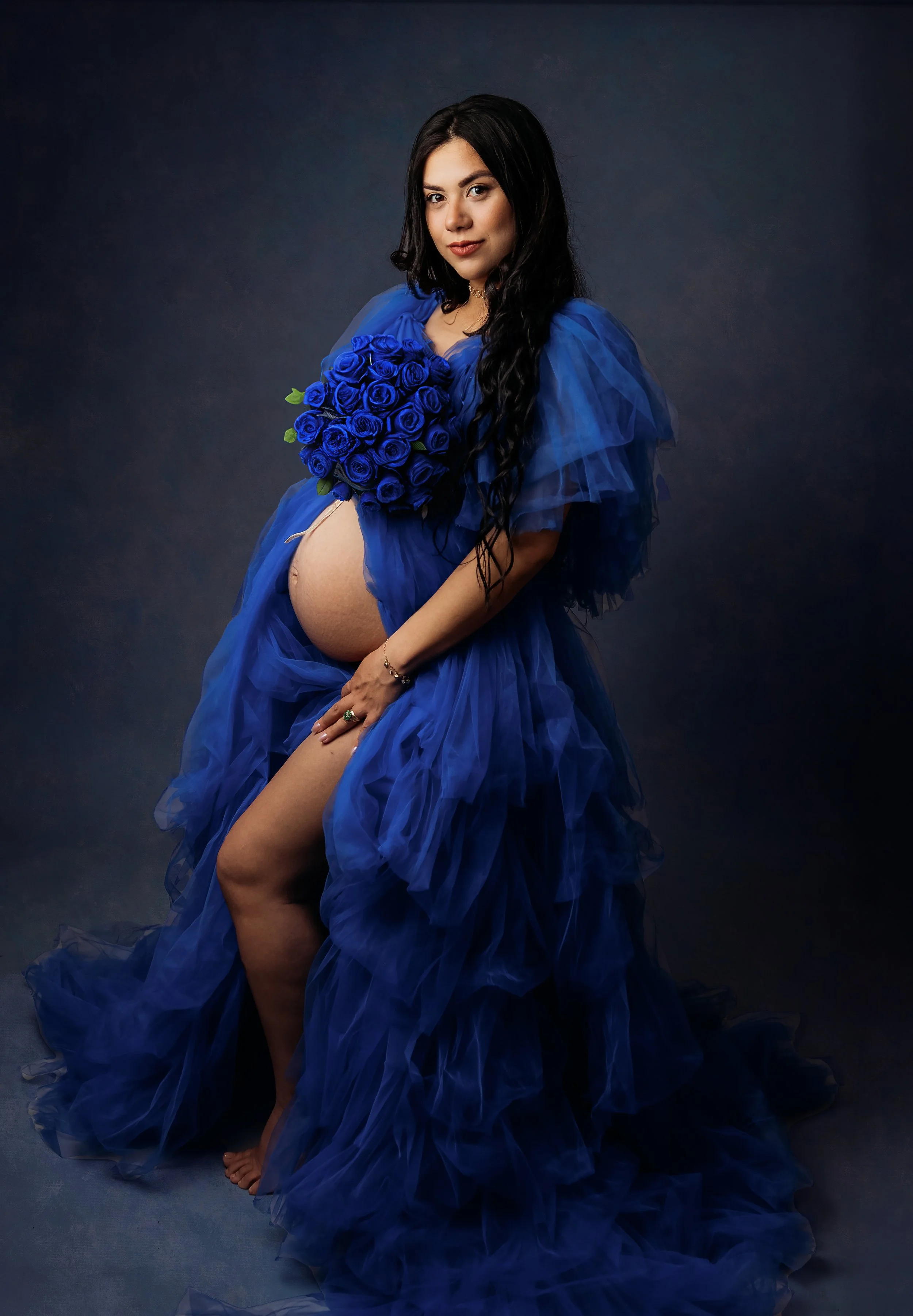 a beautiful pregnant woman with black hair facing the camera holding a dark blue rose bouquet. wearing a dark blue gown. on a dark blue textured backdrop