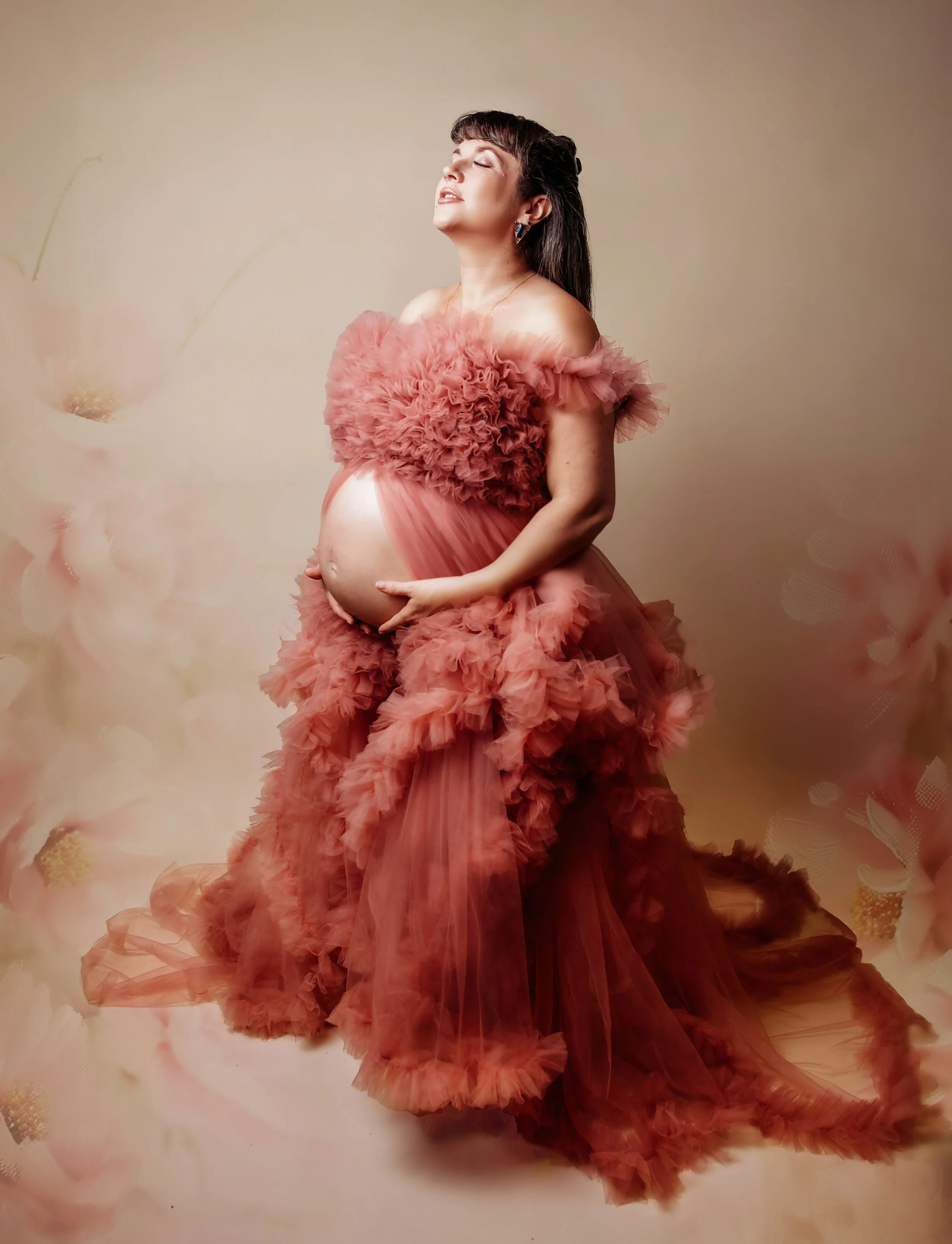 a beautiful pregnant woman with black hair wearing a floral headband. facing away from the camera with her eyes gently shut. holding her bump. wearing a dark pink. on a floral beige backdrop with a larger panning showing more of the beautiful dress