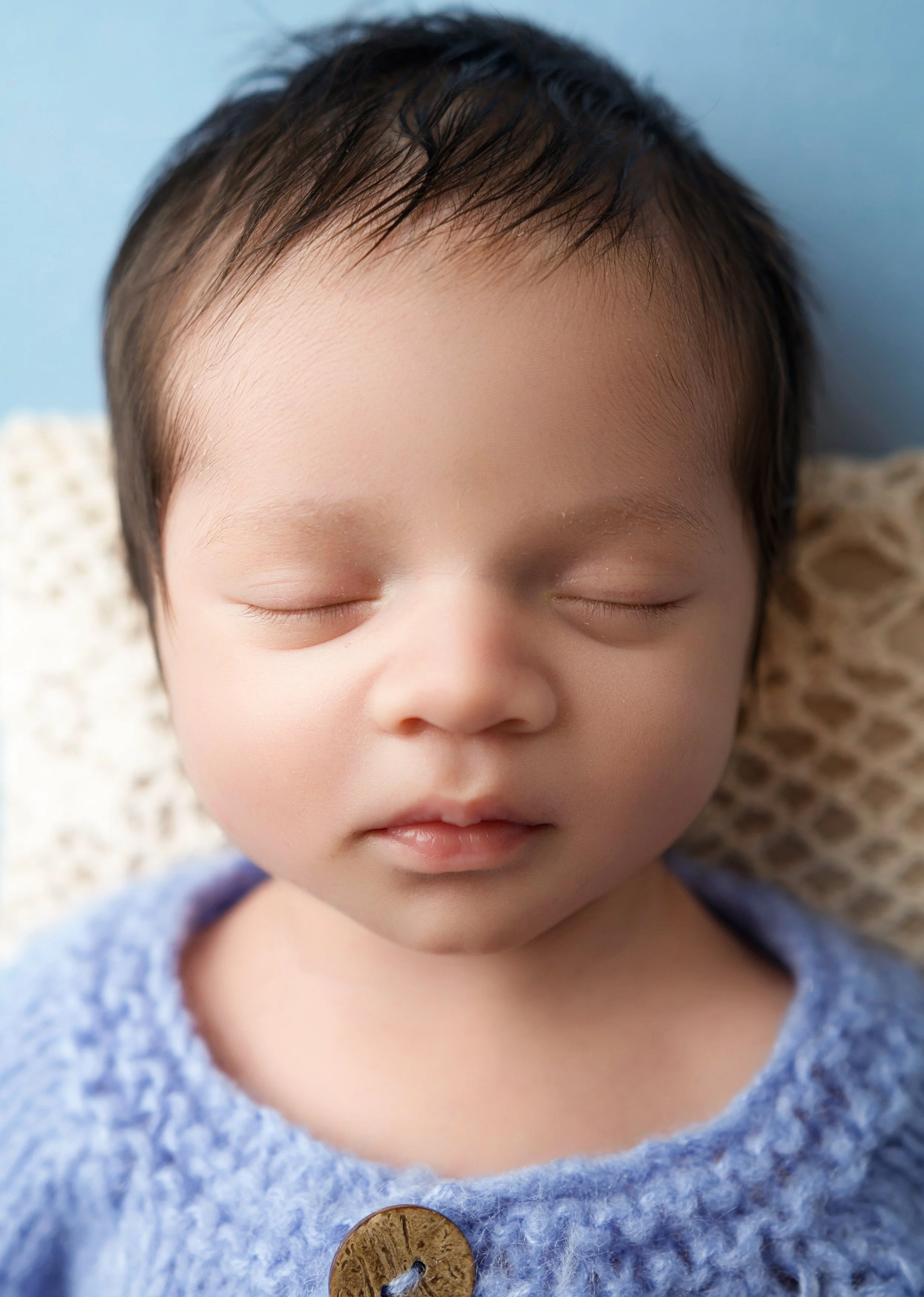 Close-up of a sleeping baby with closed eyes, dark hair, wearing a blue knitted sweater with a wooden button, lying on a beige textured blanket against a blue background.