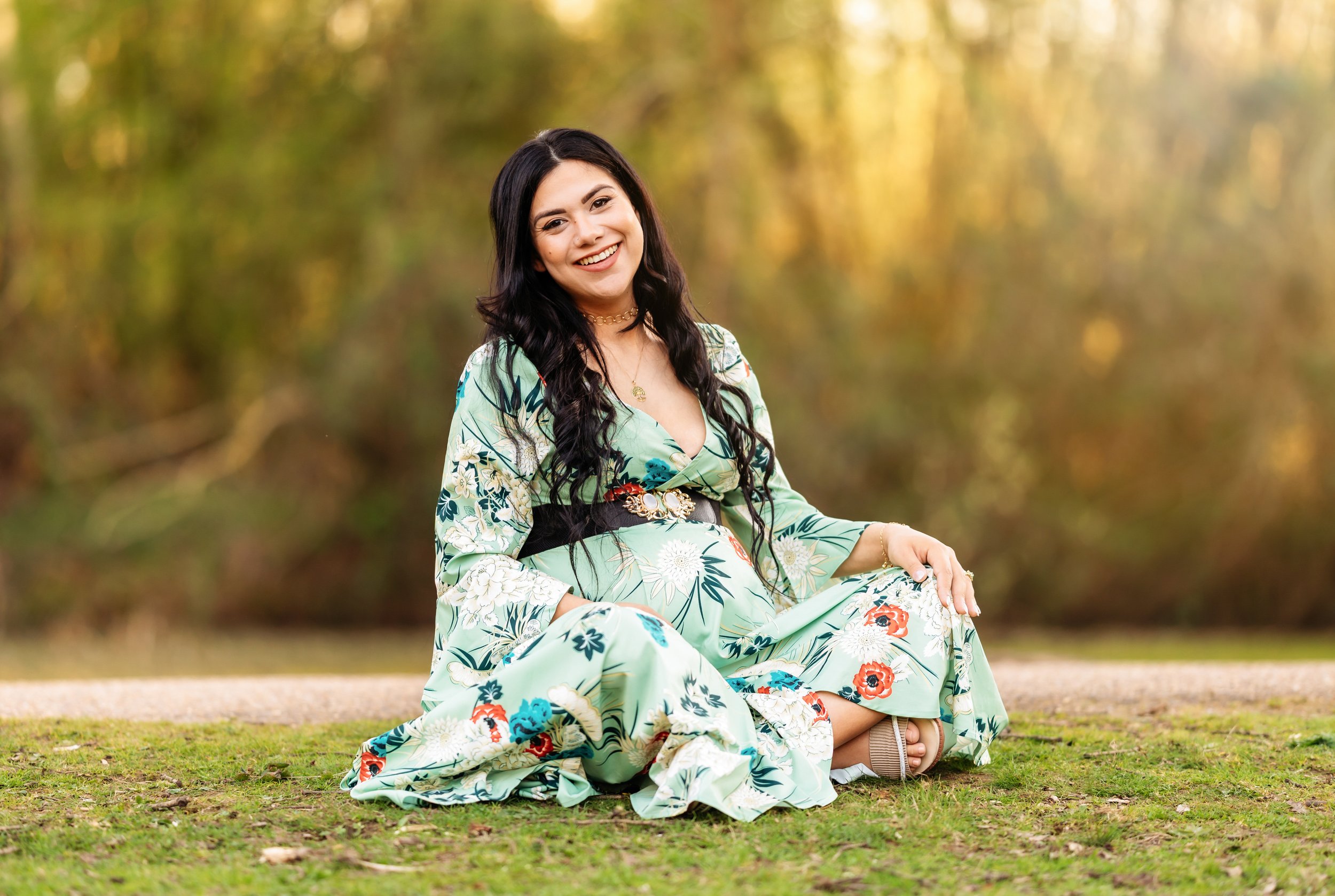 A woman sitting on grass outdoors, wearing a floral dress with long dark hair, smiling at the camera, with a blurred background of trees and autumn foliage.