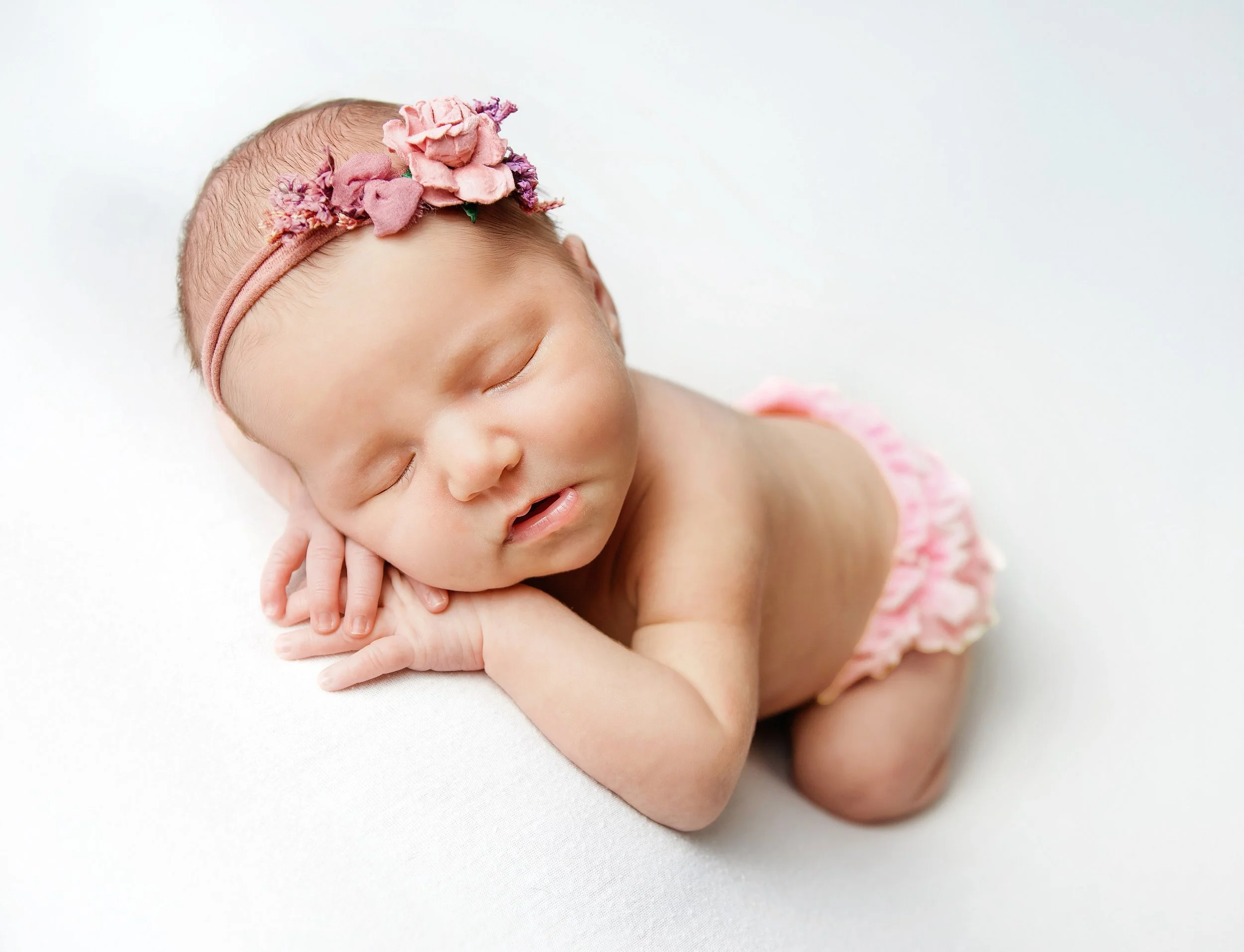 A newborn baby peacefully sleeping on a white surface, wearing a pink floral headband and ruffled pink diaper.