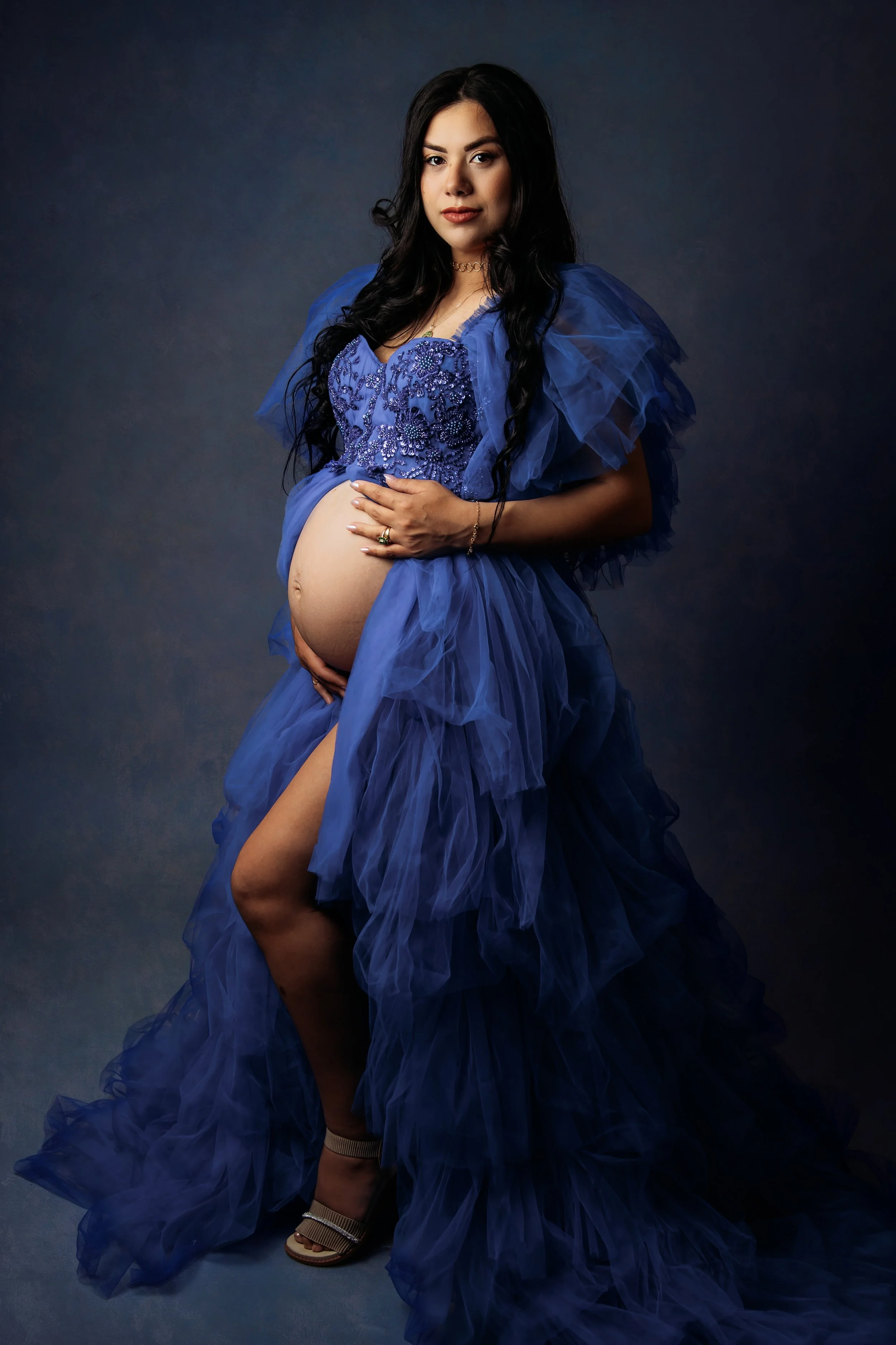 a beautiful pregnant woman with black hair facing the camera holding a her bump wearing a dark blue gown. on a dark blue textured backdrop