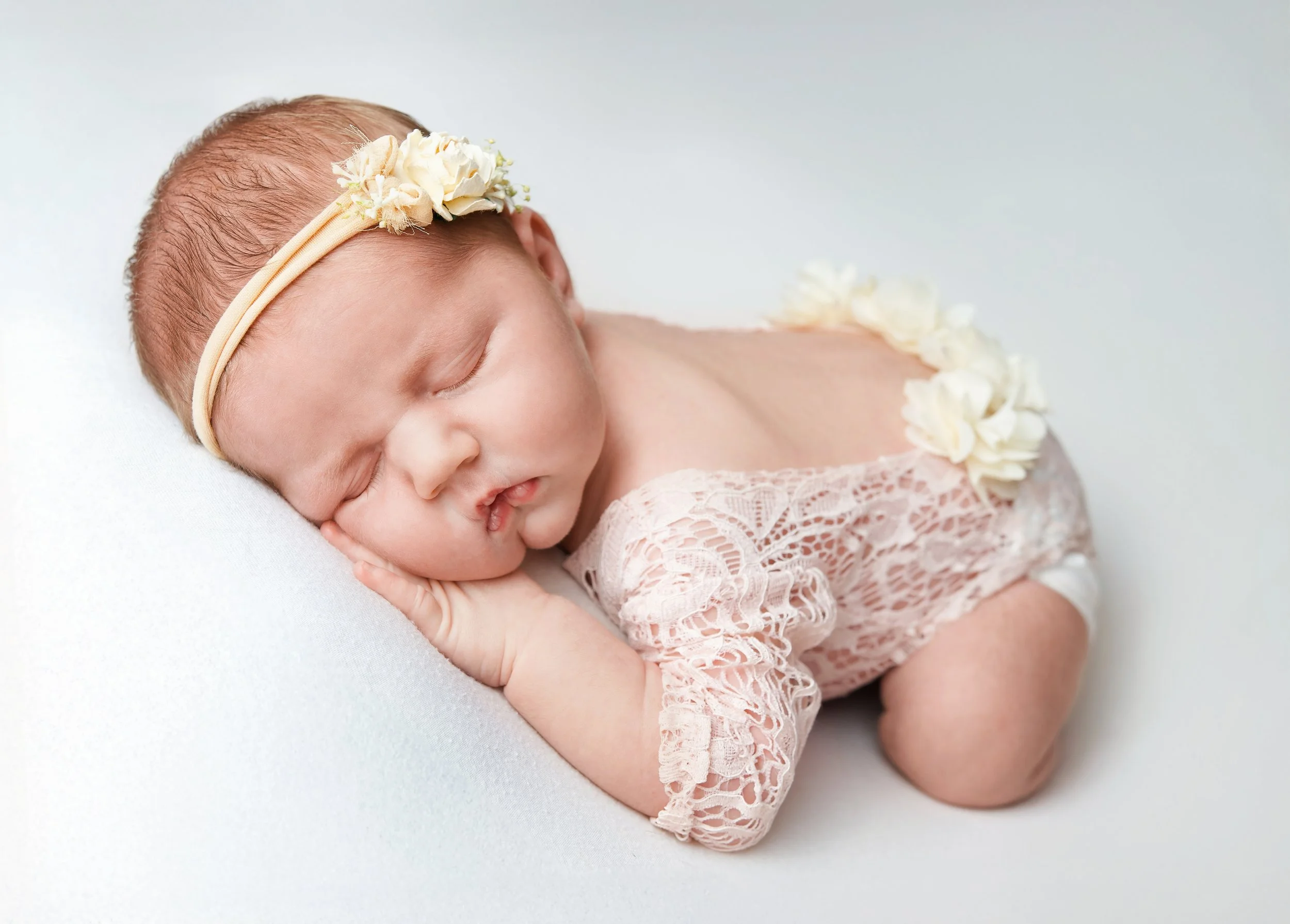 newborn girl with floral headband and floral outfit laid with hand on cheek in a white background , newborn studio photoshoot