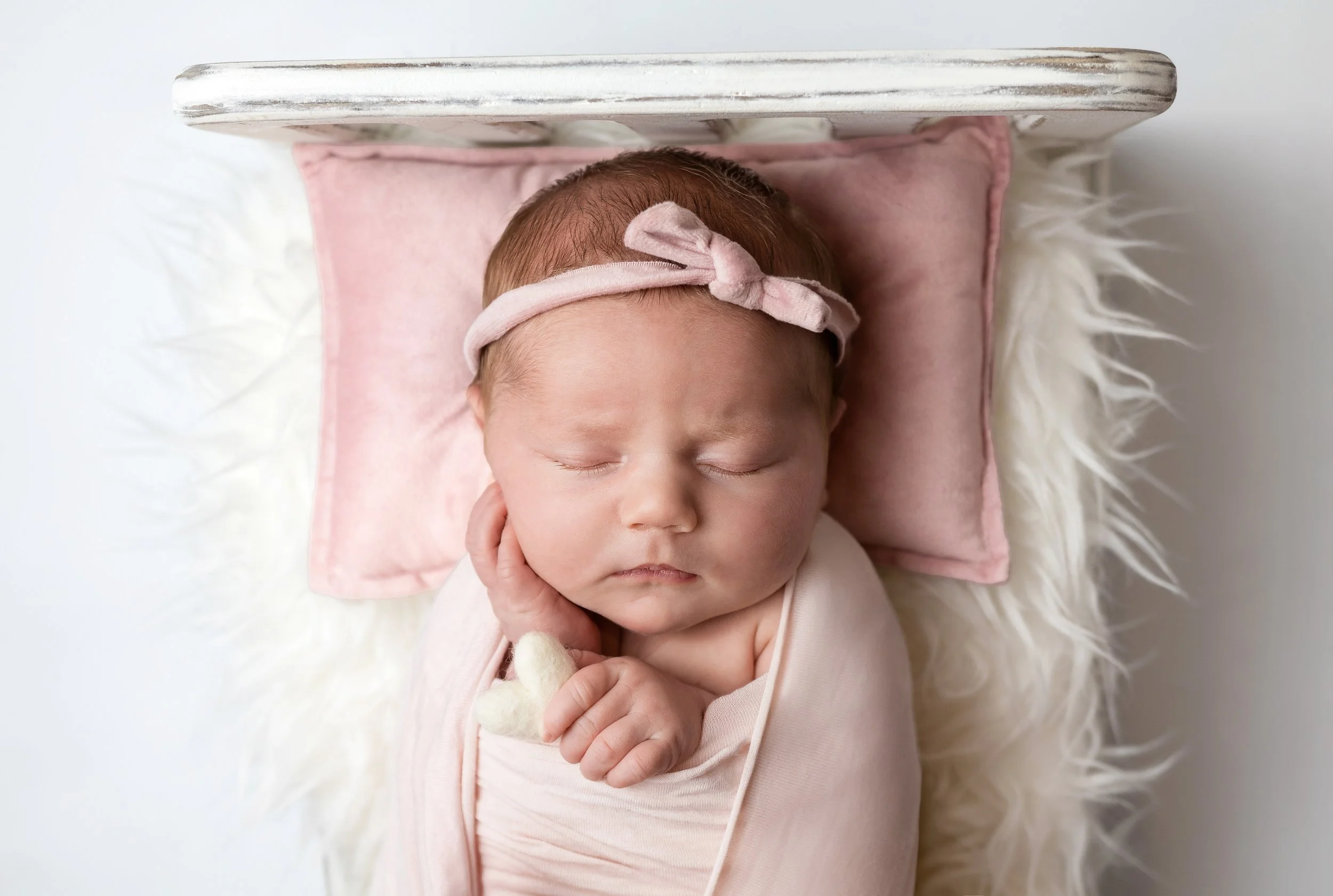 newborn girl laid on a bed with a fluffy white blanket with head laid on a pink pillow. she is holding a white felt heart and a pink headband on a white backdrop newborn studio photoshoot