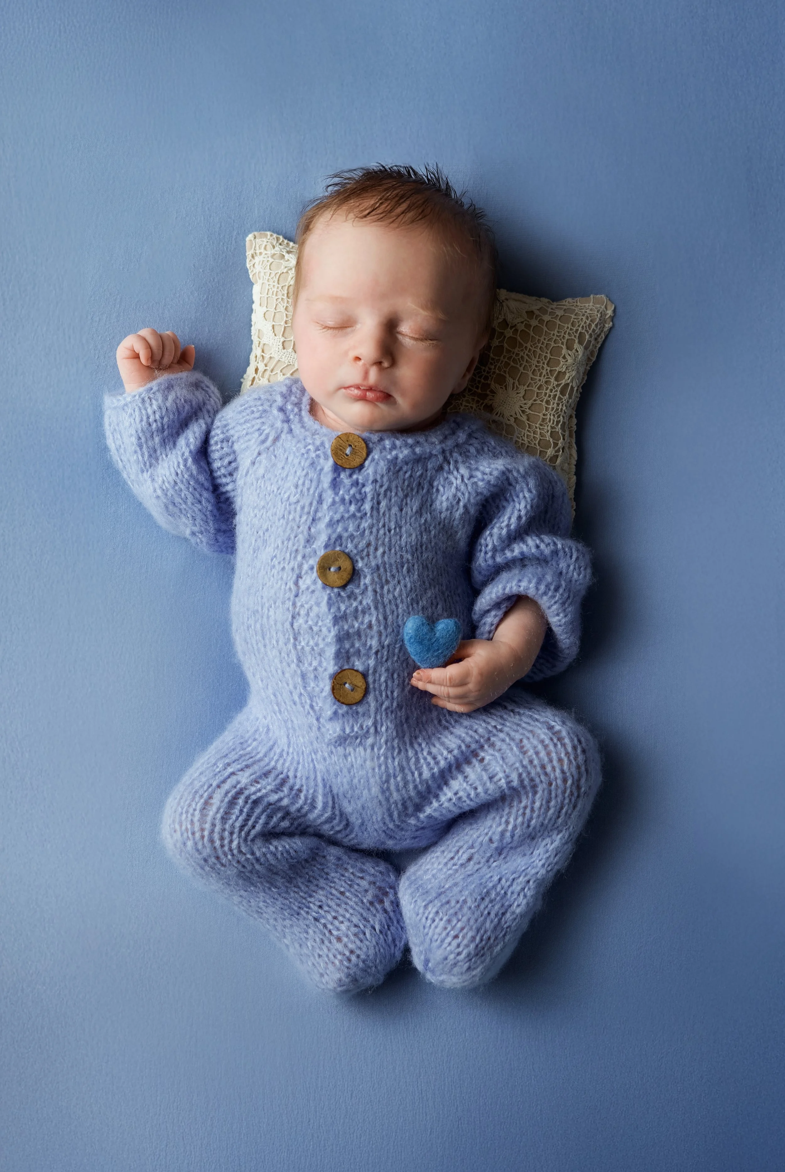 newborn boy laying on his back for photoshoot. he is wearing a blue knitted outfit and holding a blue felted heart on a blue background