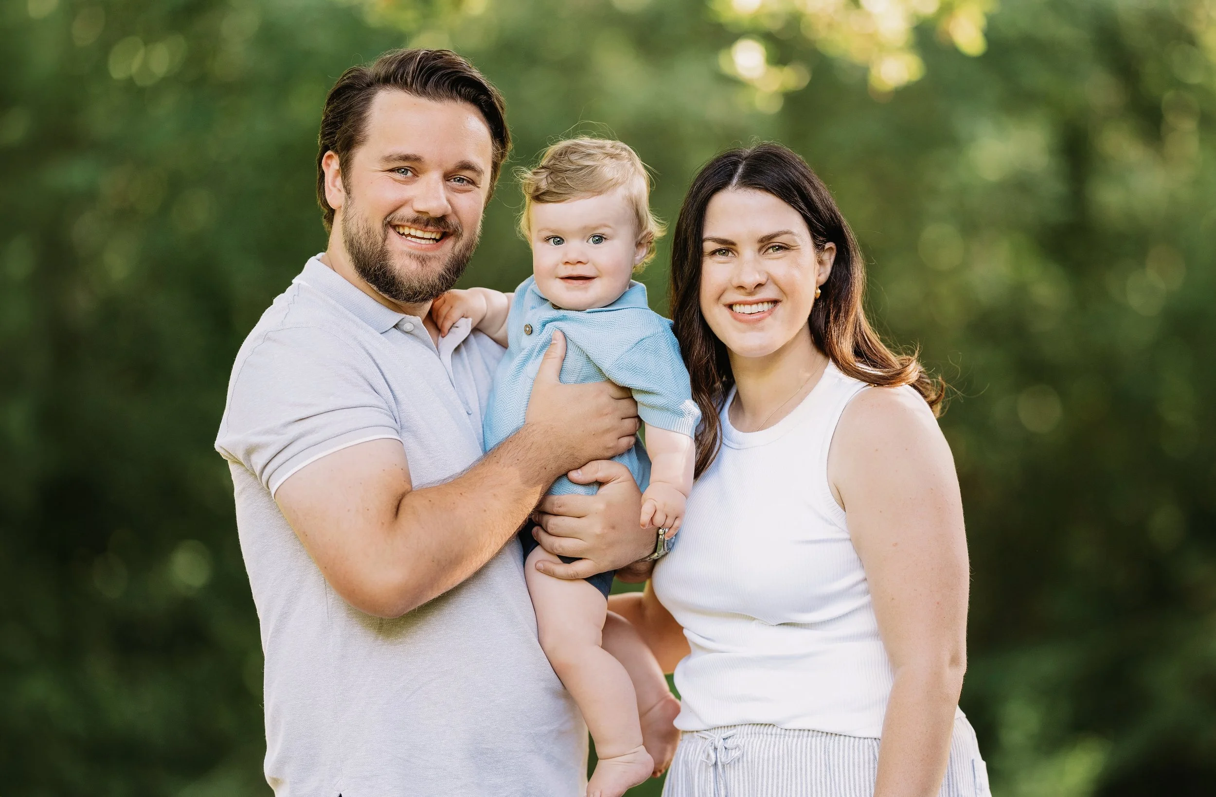 a portrait of a family of 3 wearing light colors with the background blurred. all looking at the camera and smiling