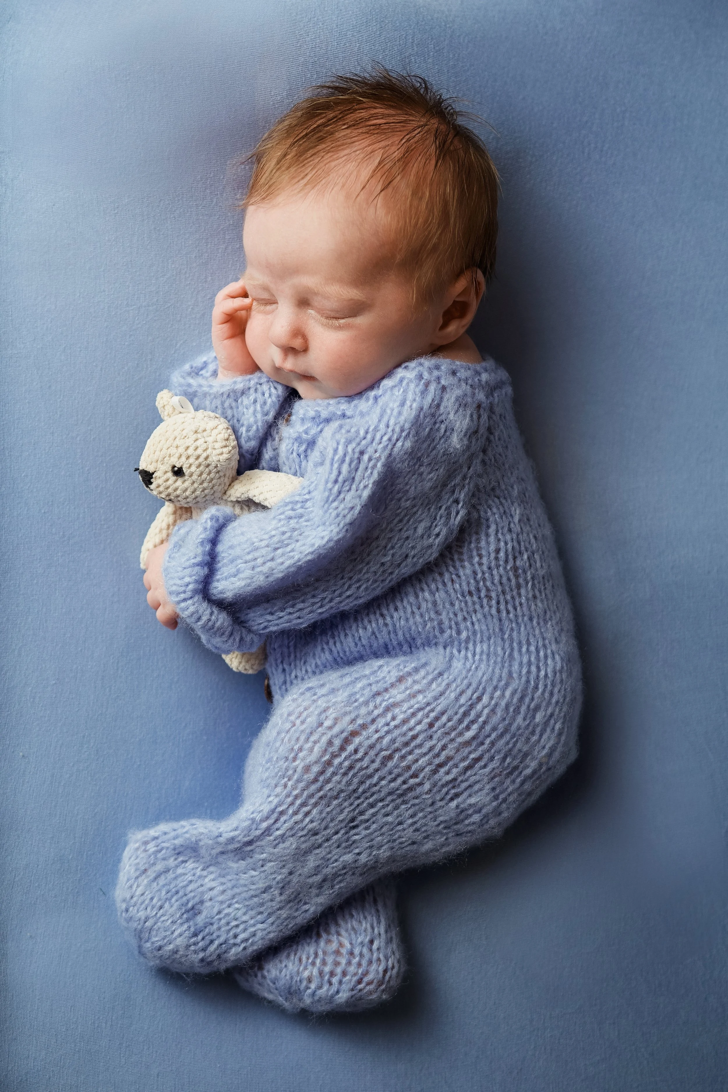 Newborn boy in a blue knitted outfit laid on his side on a blue backdrop cuddling a teddybear