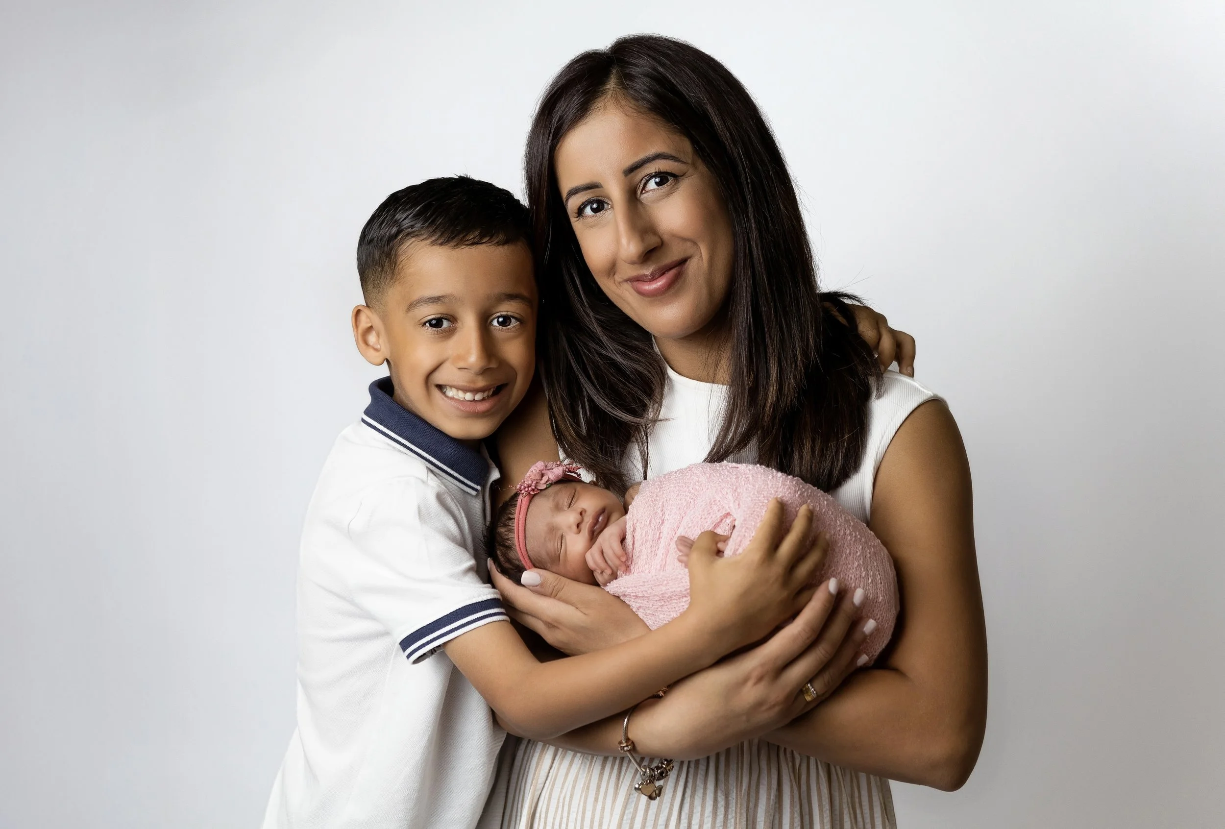 studio photo on white background of mum cradling newborn girl in her arms wearing a pink wrap and pink headband. with brother cuddling mum and baby. all looking at the camera and smiling. mum and son wearing white