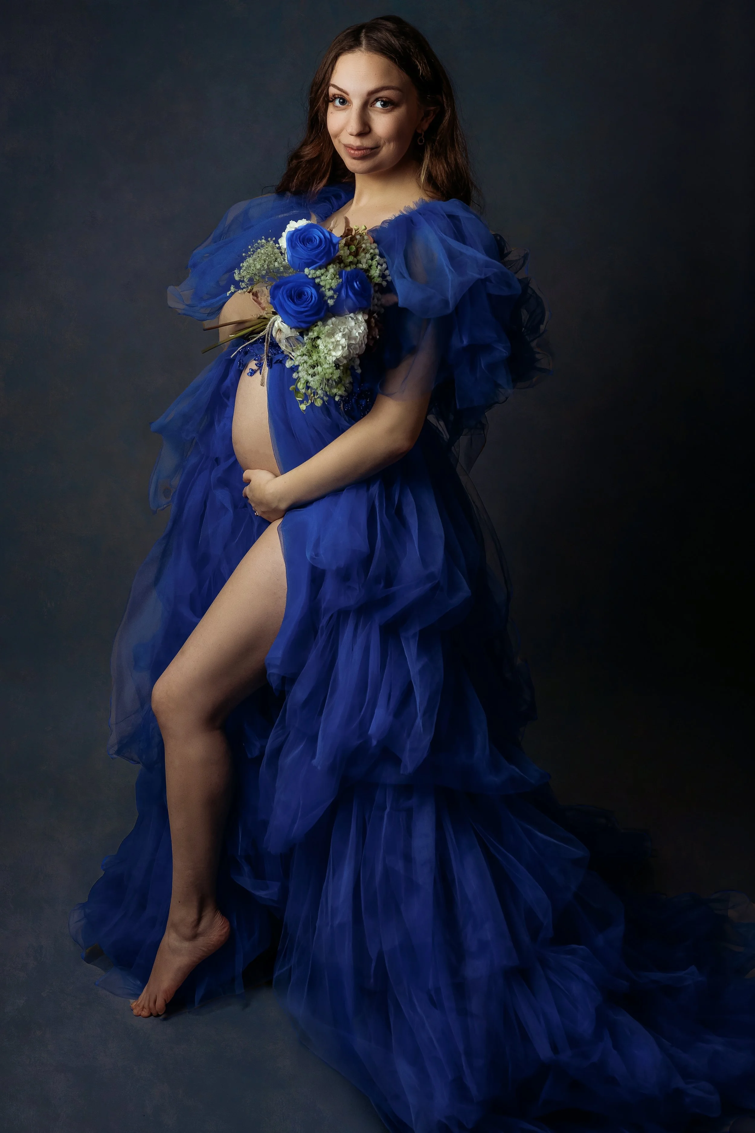 a beautiful pregnant woman with brown hair facing the camera smiling holding a dark blue rose bouquet. wearing a dark blue gown. on a dark blue textured backdrop