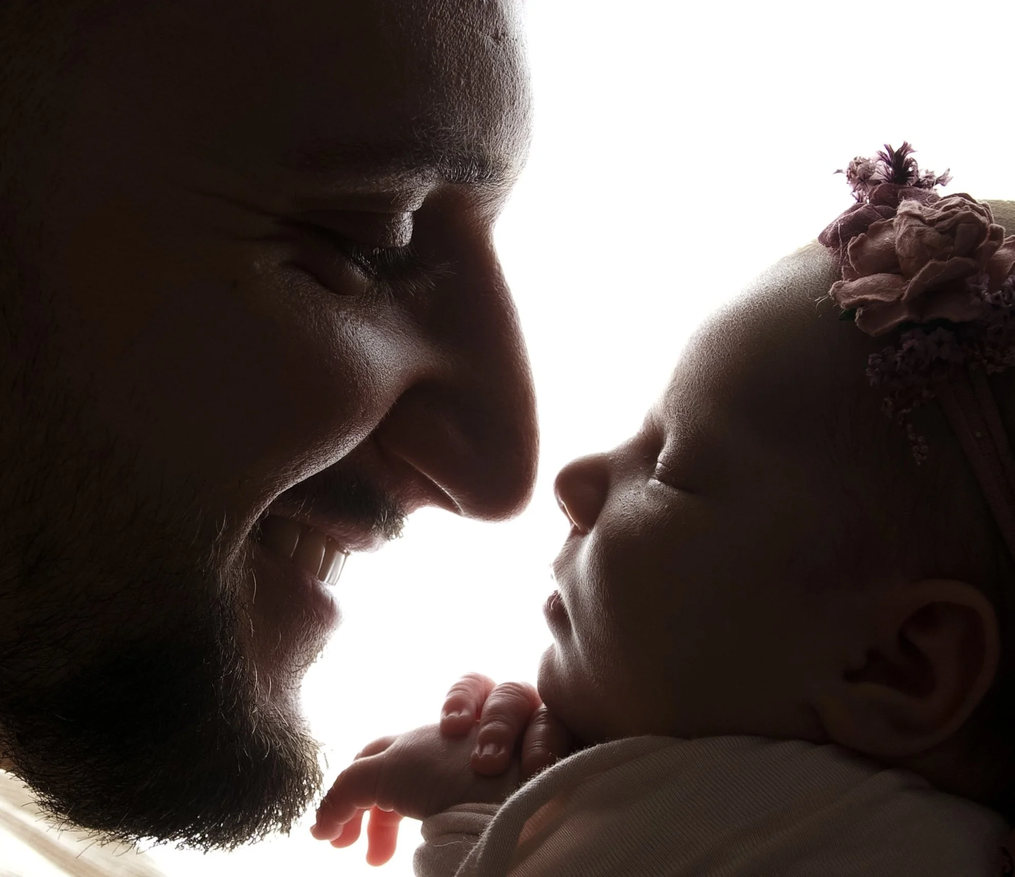 A touching moment between a father and his infant daughter, sharing a gentle smile as their foreheads touch, backlit with a bright light.
