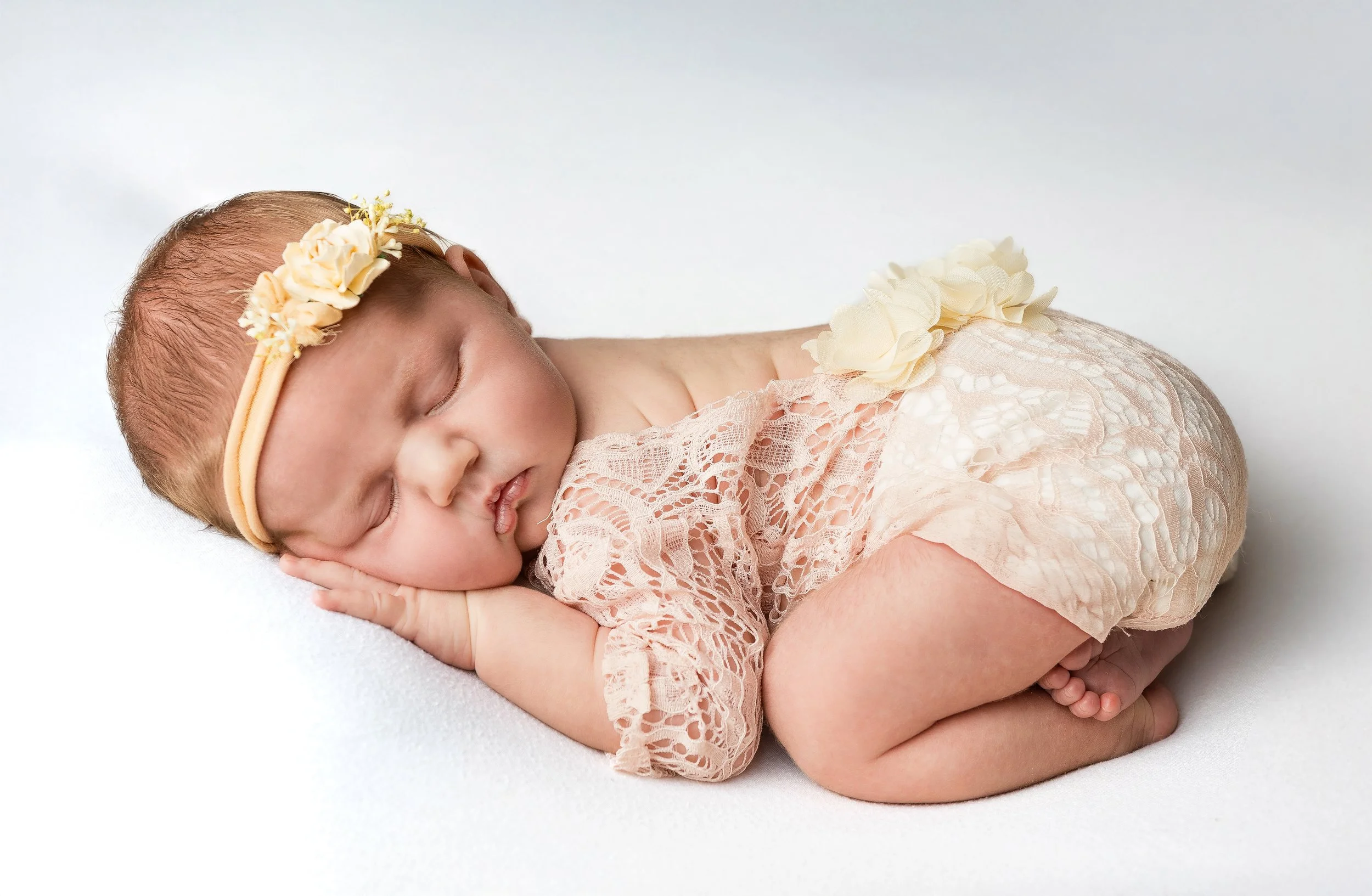 A sleeping newborn baby girl lying on her side, wearing a lace pink outfit with yellow floral accents and a matching headband with flowers, on a white surface.
