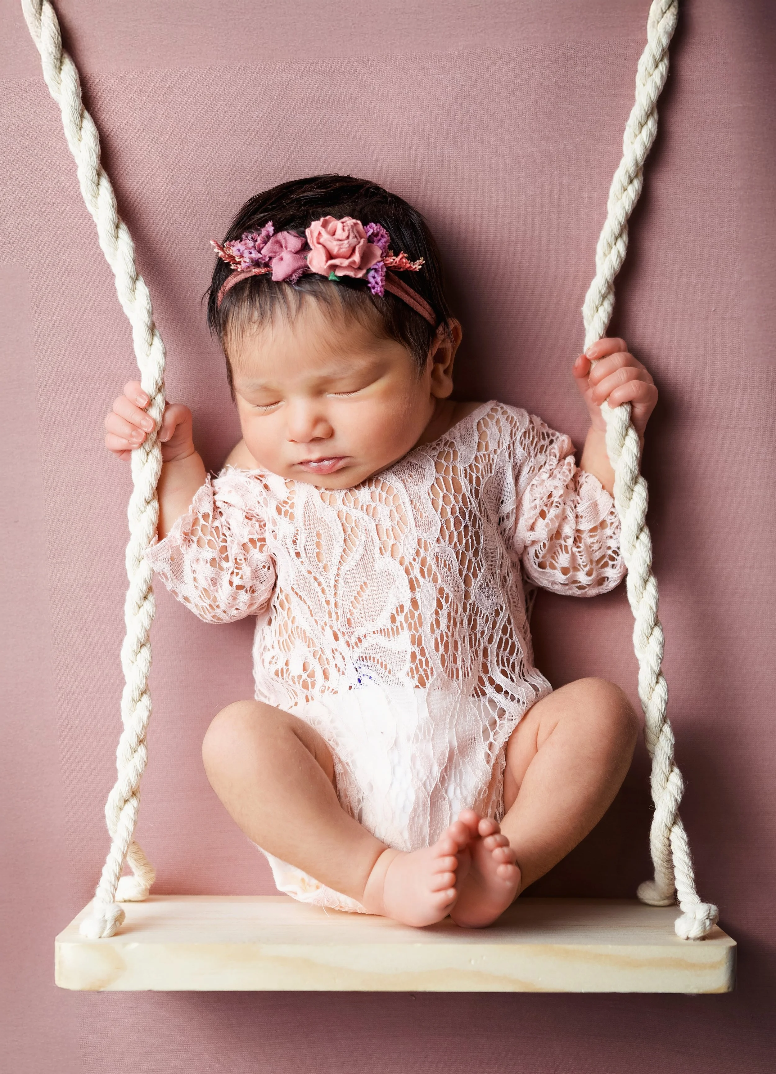 baby in a pink floral outfit with a pink floral headband laying on a swing looking like she's swinging holding onto the handles. there is a pink background