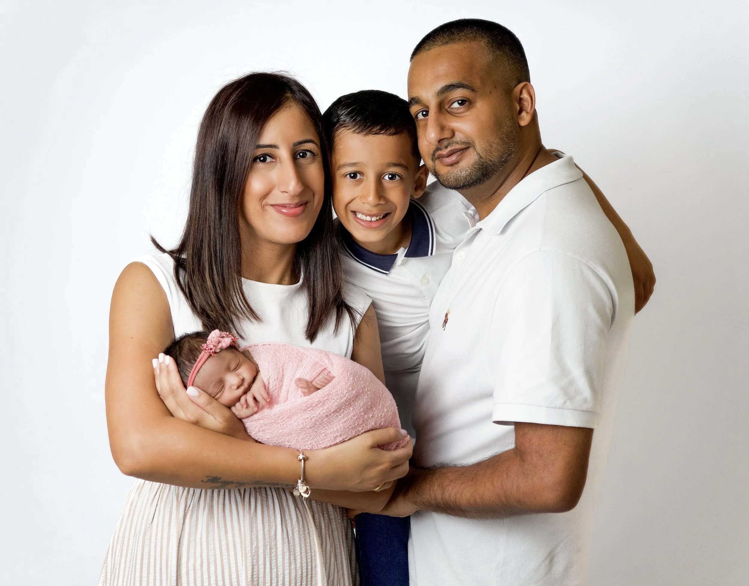 studio photo of a family of 4. all are wearing white except newborn baby girl which is in a pink wrap with a pink headband. mum is cradling baby In her arms. boy with his head between his parents smiling