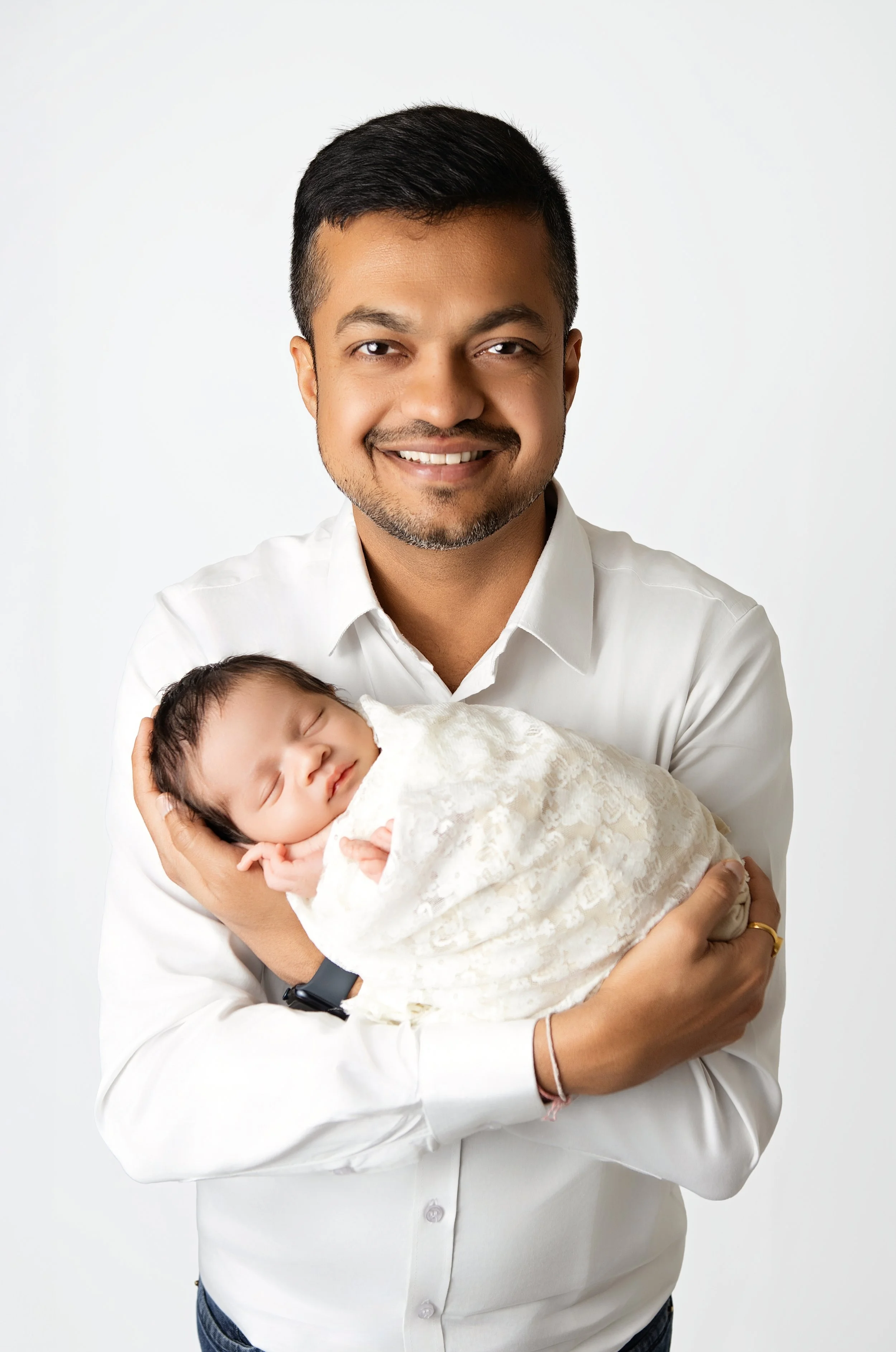 A man with short black hair, a beard, and a white shirt smiling while holding a sleeping baby wrapped in a white blanket against a plain white background.