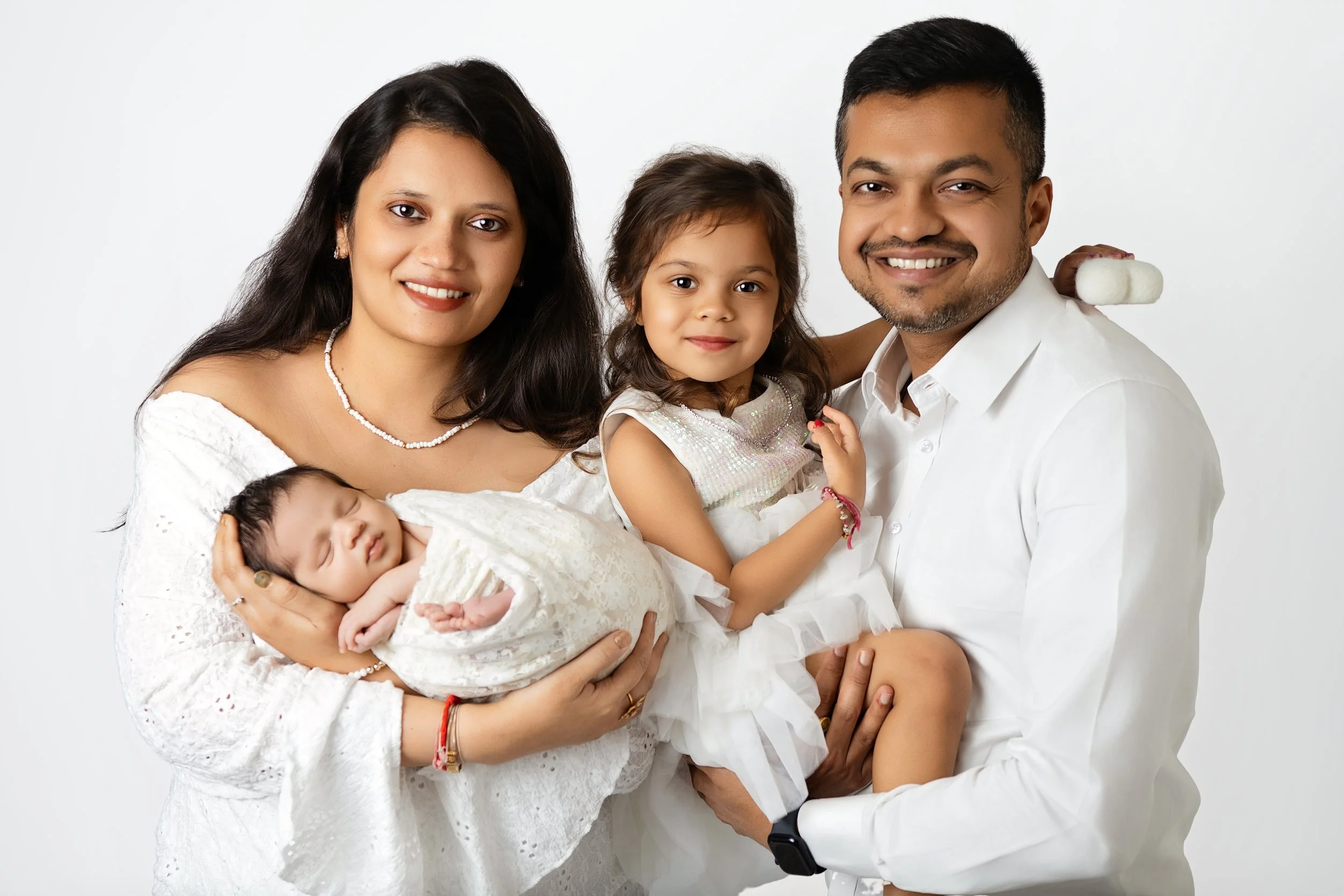 A smiling Indian family of four, with a mother holding a newborn baby, a father holding a young girl, all dressed in white against a white background.