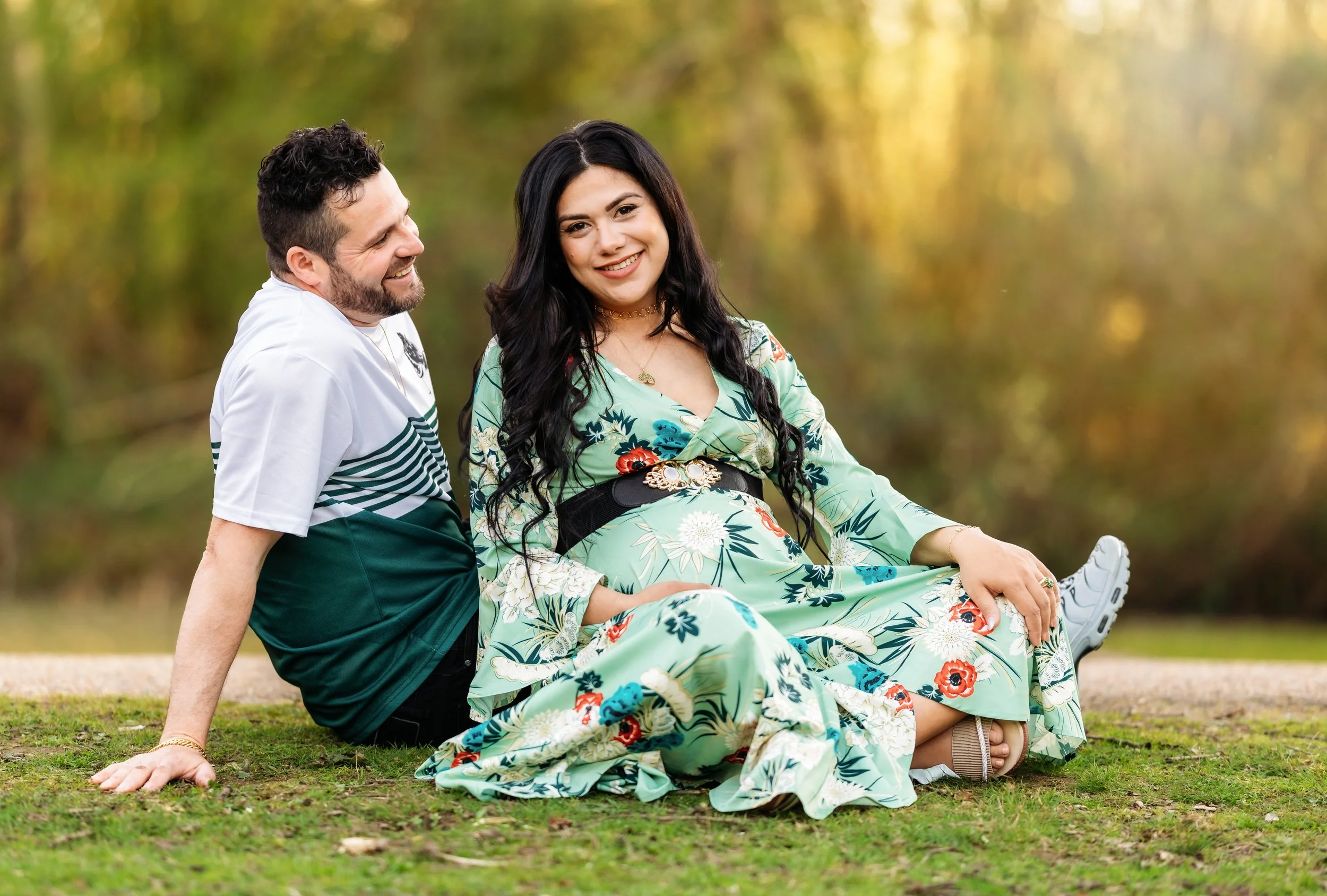 maternity photoshoot of a pregnant lady in a floral dress with her husband next to her in an outdoor setting sitting down on the ground with her legs crossed and her hands on her knees . her husband is sitting on his side smiling at her