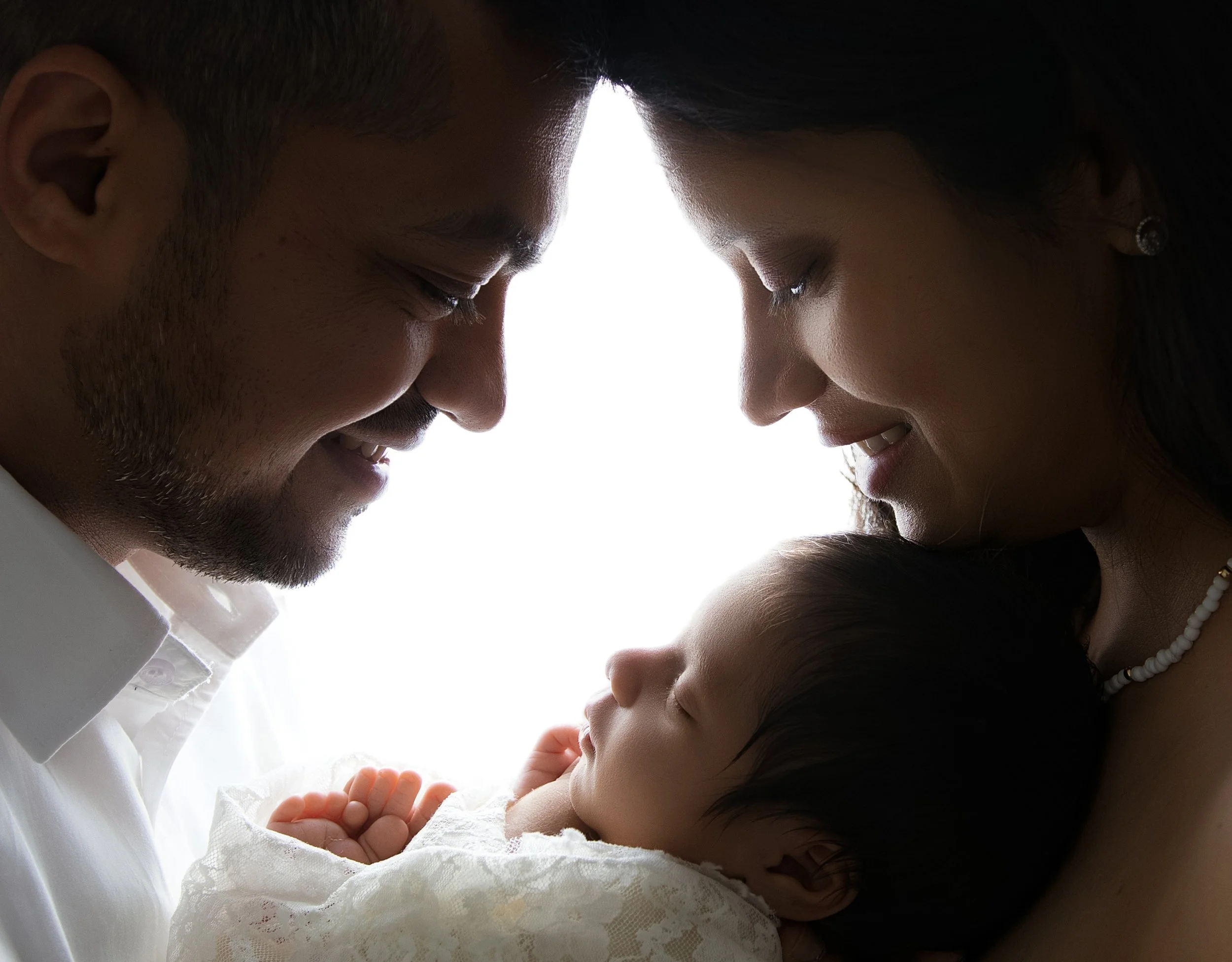 A family of three, a man, a woman, and a baby, with their foreheads touching, creating a silhouette against a bright background.