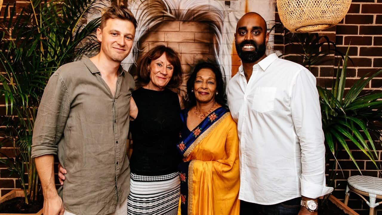 Group of five diverse people standing together indoors, smiling for the camera, with plants and brick wall background.