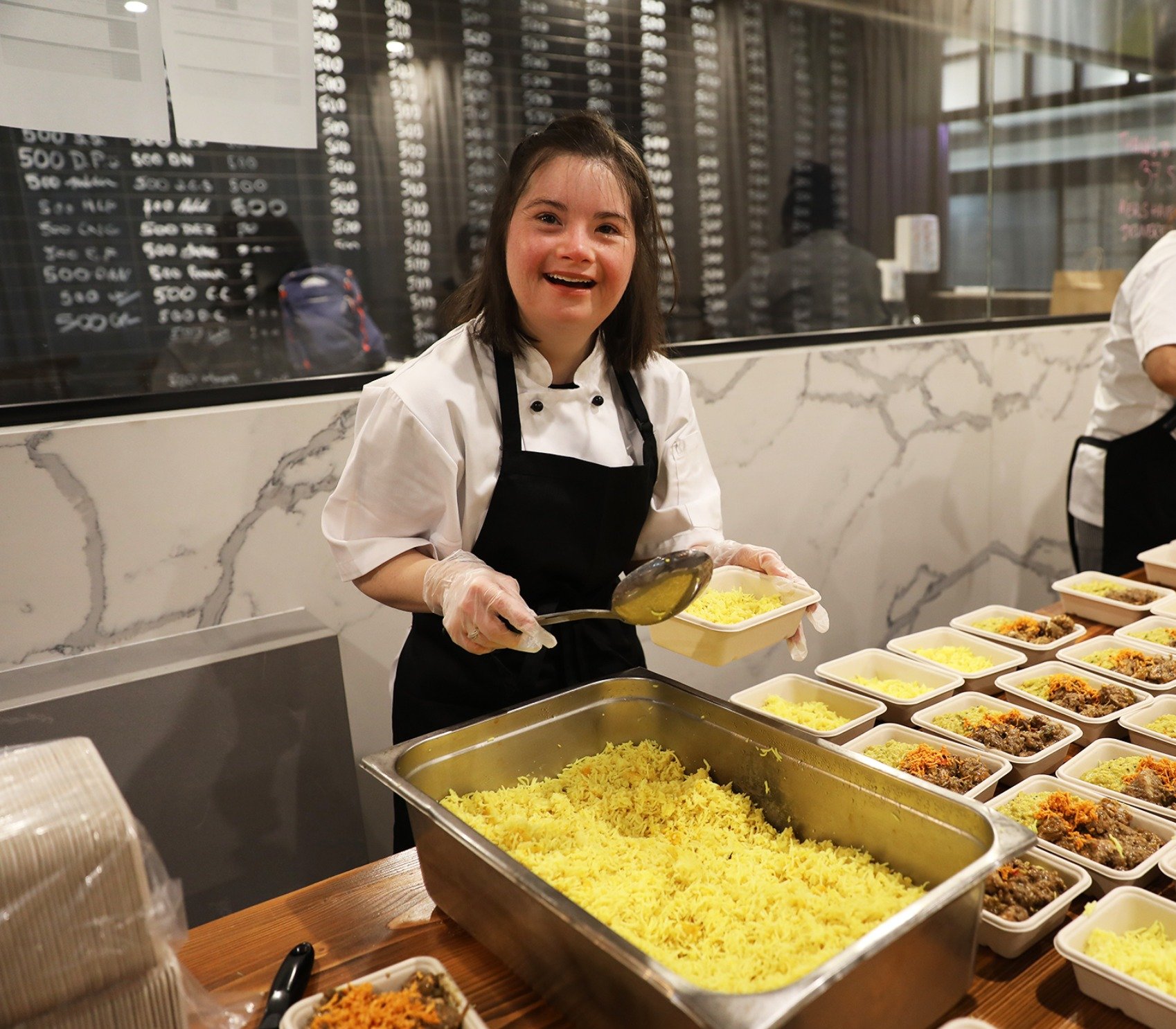 A smiling woman in a chef's uniform with a black apron serving yellow rice from a large tray into takeaway containers at a food service station.