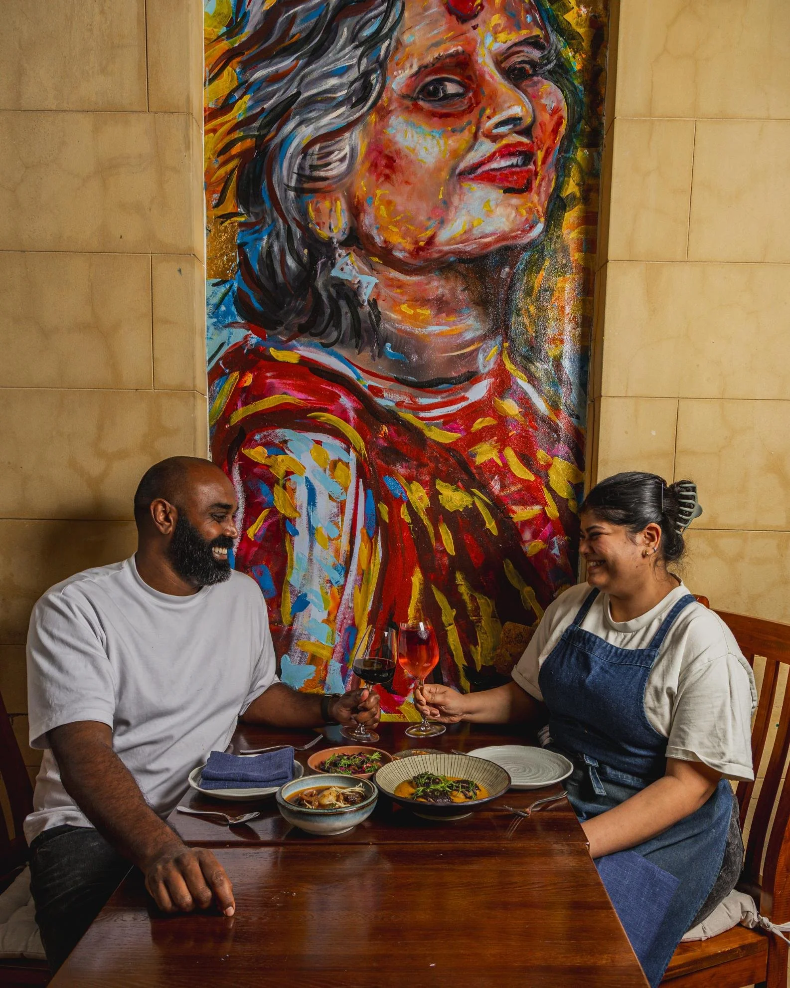 A man and a woman sharing a toast with glasses of red wine in a restaurant. The man has a beard and is wearing a white t-shirt, the woman has dark hair pulled back and is wearing a light-colored shirt with a blue apron. They are sitting at a wooden t