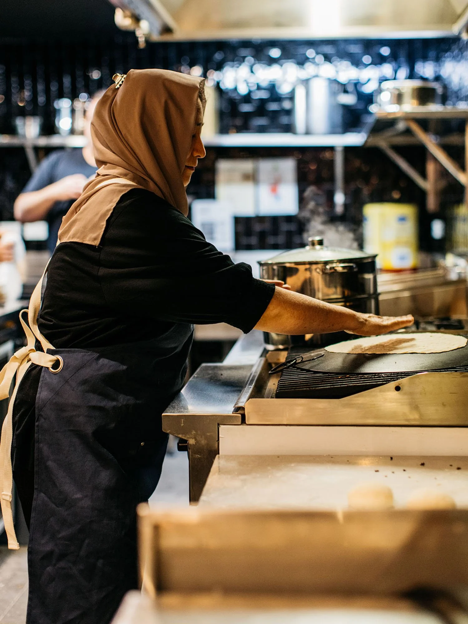 A woman wearing a beige hijab and black apron preparing flatbread in a commercial kitchen.