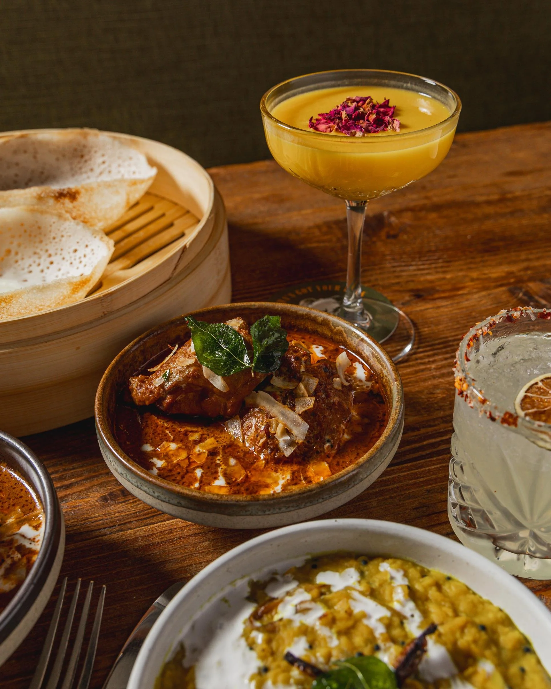 Assorted Indian dishes on a wooden table, including naan bread, a spicy meat curry, yellow dessert garnished with dried rose petals, and a glass of Indian-style cocktail with lemon slice and rim salt.