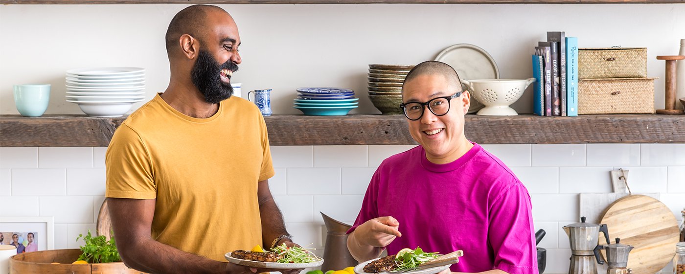 Two men smiling and holding plates of food in a kitchen with dishes and books on shelves behind them.
