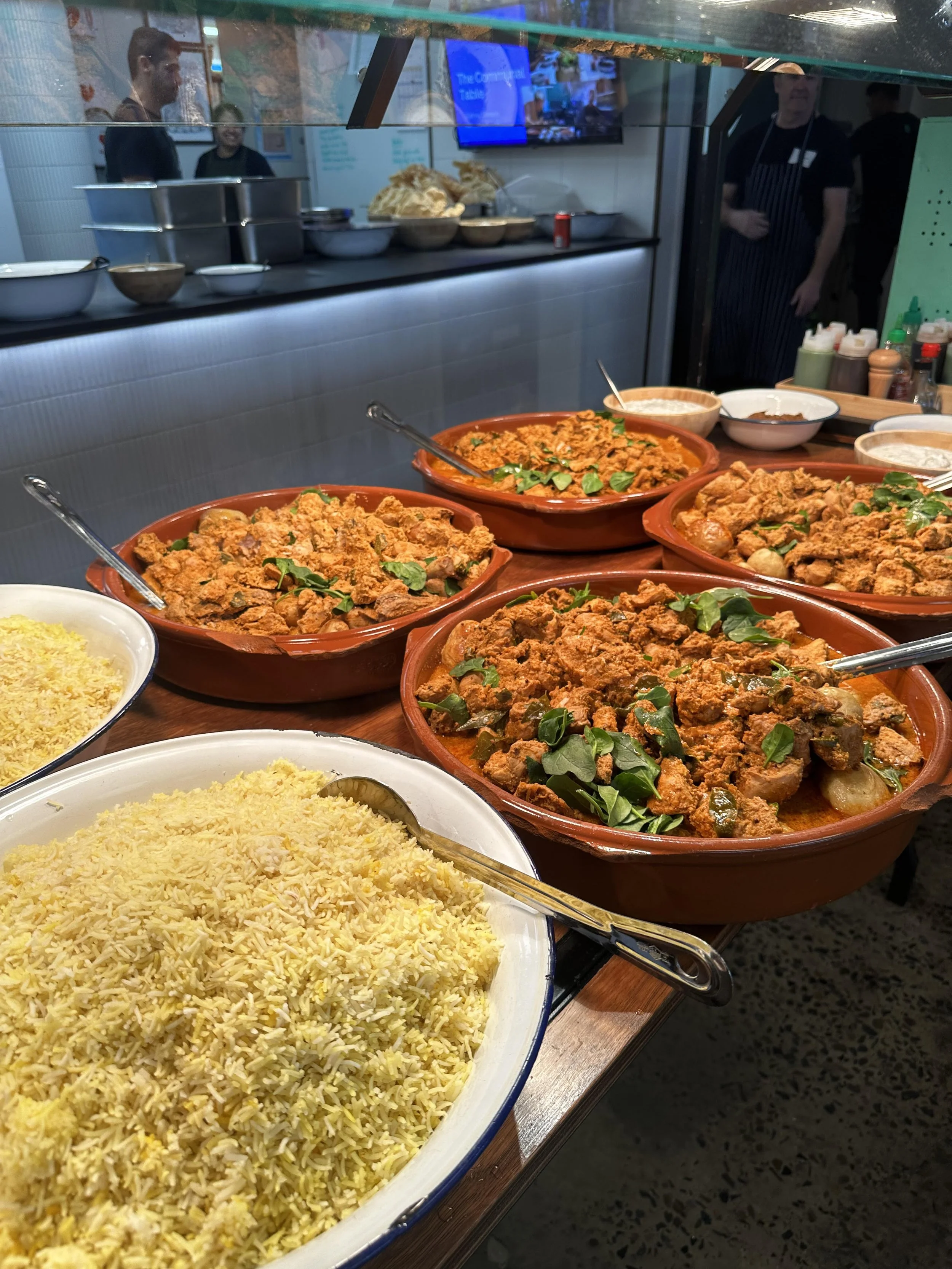 Large bowls of Indian curries garnished with fresh green herbs, alongside bowls of yellow rice, arranged on a buffet table at a restaurant.
