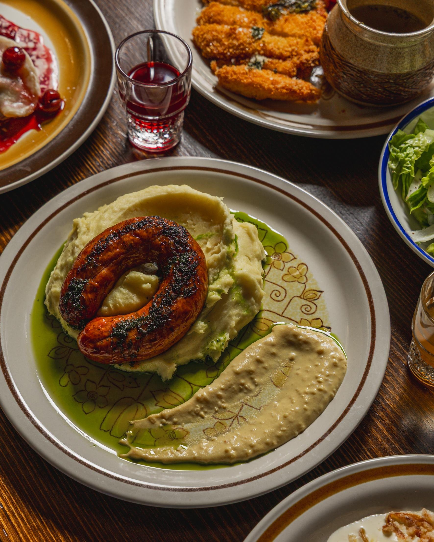 Plates of Middle Eastern dishes including grilled sausage, mashed potatoes, hummus, salad, fried fish, and a beverage.