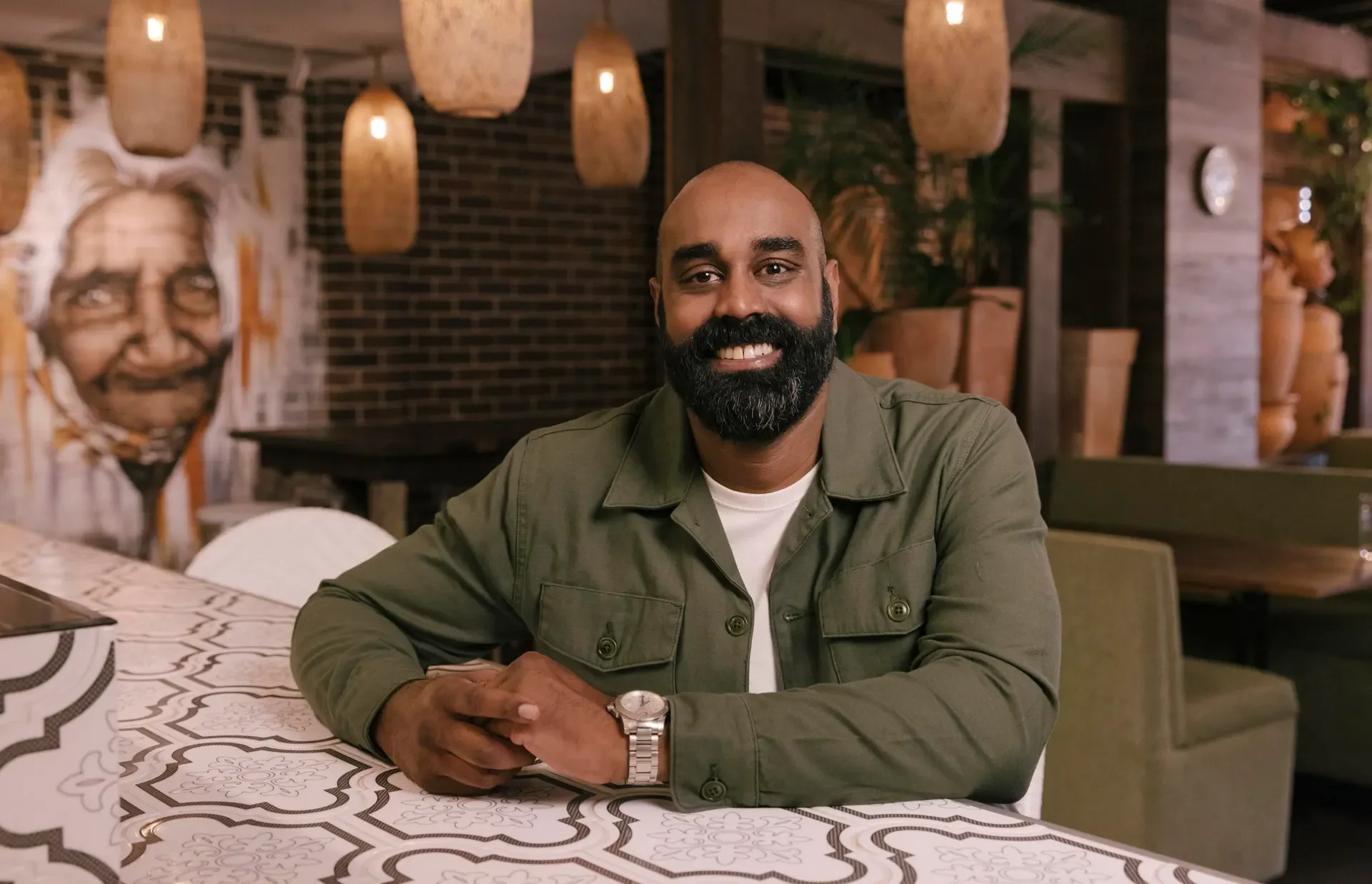 A smiling man with a beard and shaved head sitting at a table in a restaurant, wearing a green jacket, white shirt, and a watch, with decorative lighting and artwork in the background.